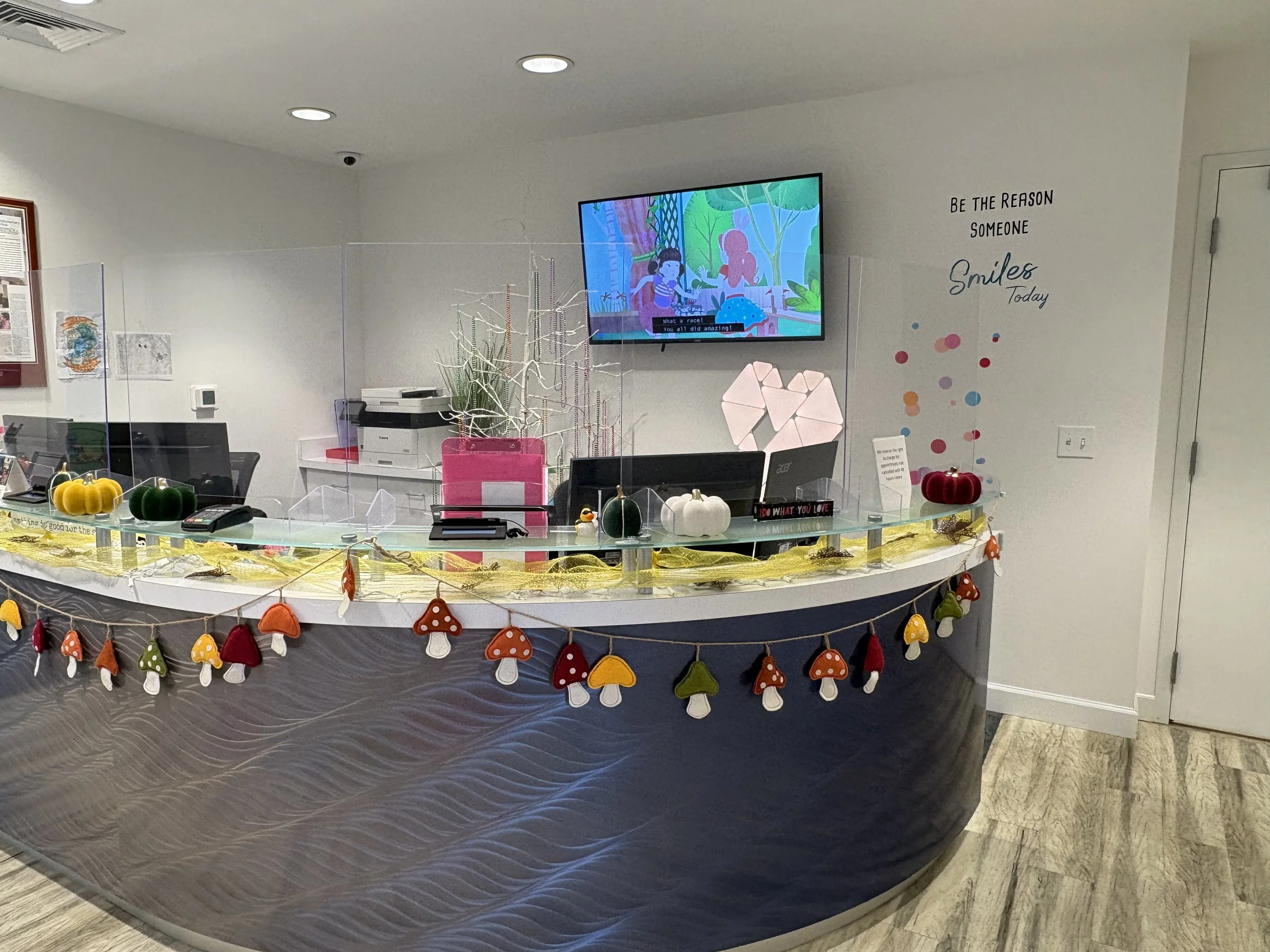 A reception desk decorated with felt mushrooms and string lights, with a computer, phones, and decorative plants on top. A wall-mounted TV displays a cartoon scene, and a motivational quote on the wall reads, 'Be the reason someone smiles today.'