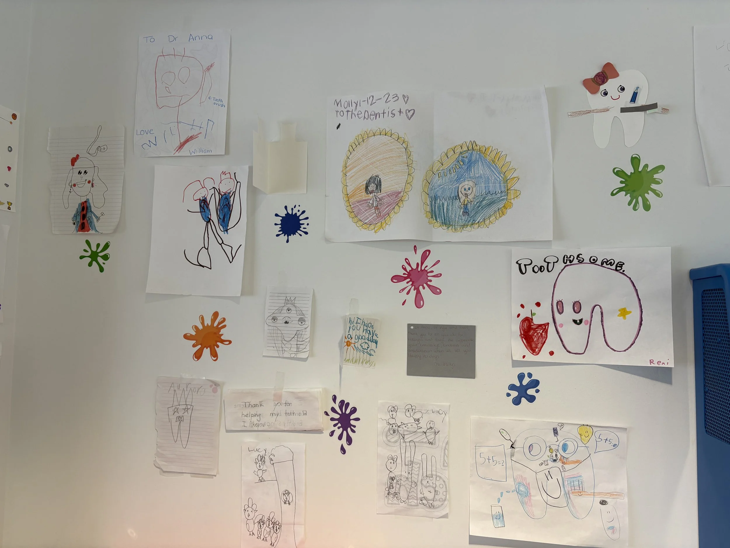 Collection of children's drawings and notes taped to a white wall, including illustrations of teeth, children, and colorful abstract shapes, with some handwritten messages and signatures.