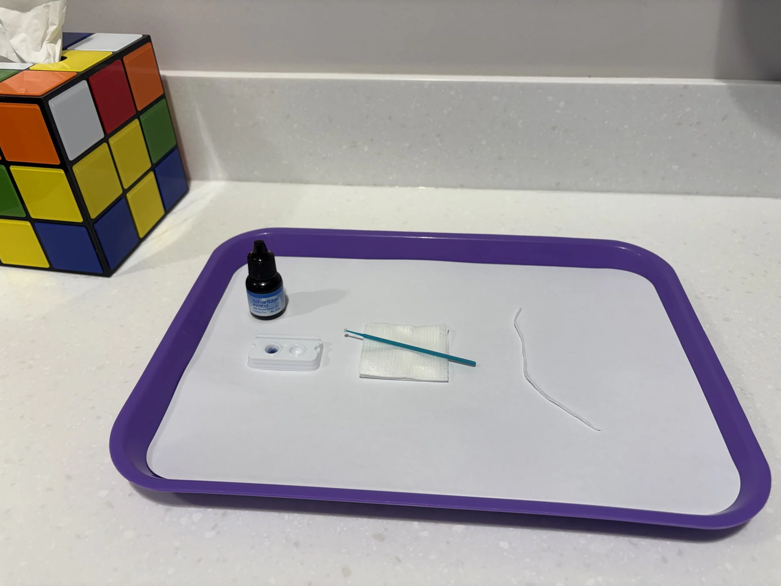 A countertop with a partially assembled purple tray, a small bottle of eye ointment, a cotton swab, a piece of gauze, and a scalpel.