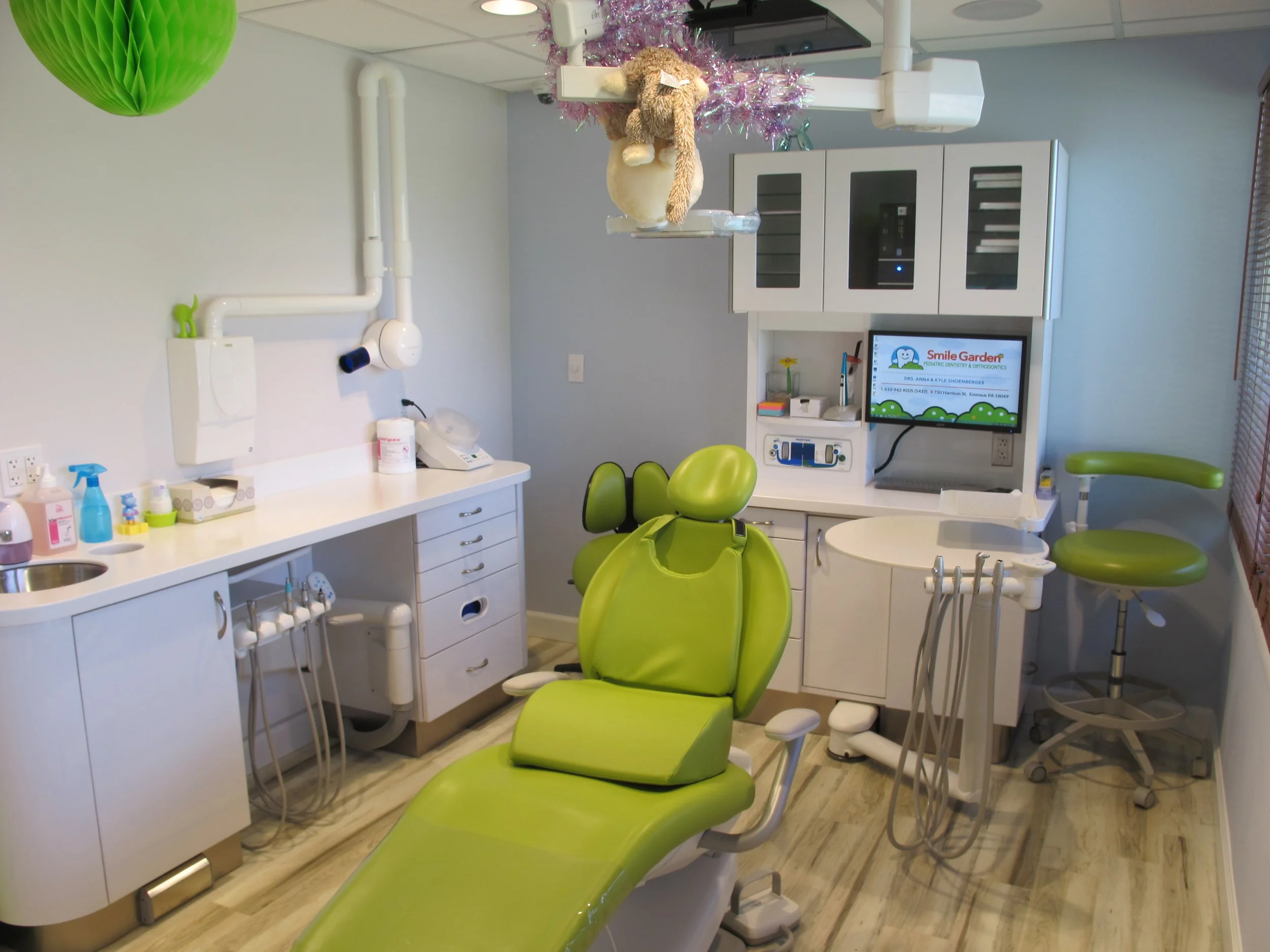 A pediatric dental clinic room with a bright green dental chair, white cabinets, a computer monitor displaying the clinic's logo 'Smile Garden Pediatrics Dentistry & Orthodontics' and a plush dog toy hanging from the ceiling decoration.