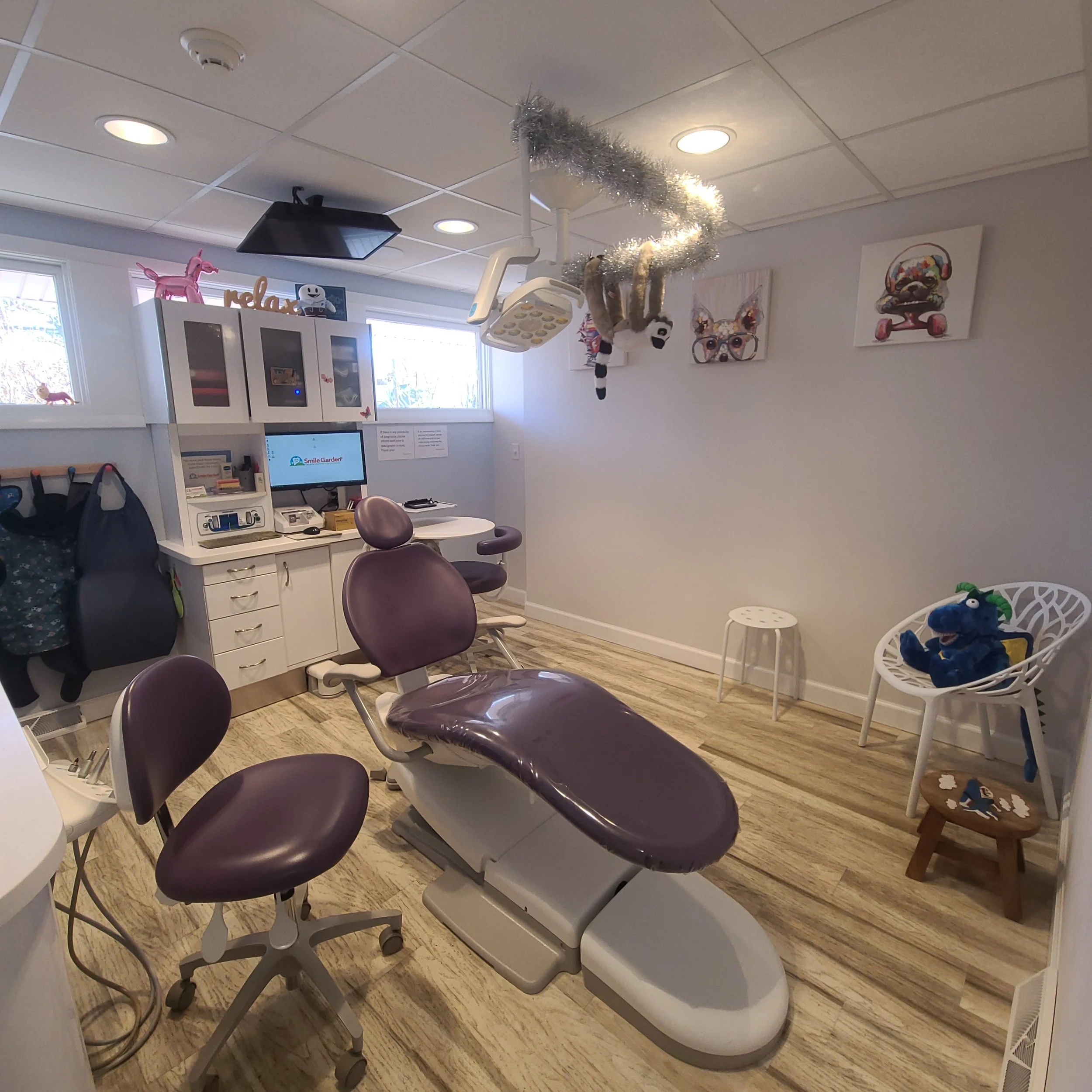 View of a dental office with dental chairs, equipment, and decorations on the wall and ceiling, including a hanging tinsel wreath and plush toys.