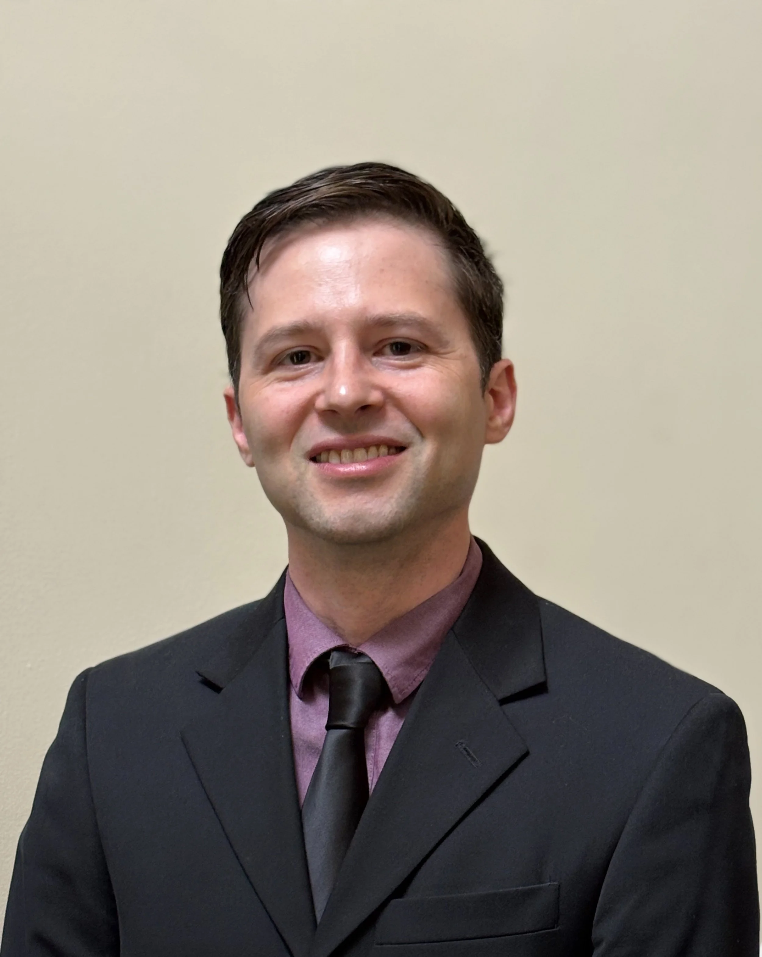A man with short brown hair and fair skin, wearing a black suit, purple shirt, and black tie, smiling in front of a plain beige wall.