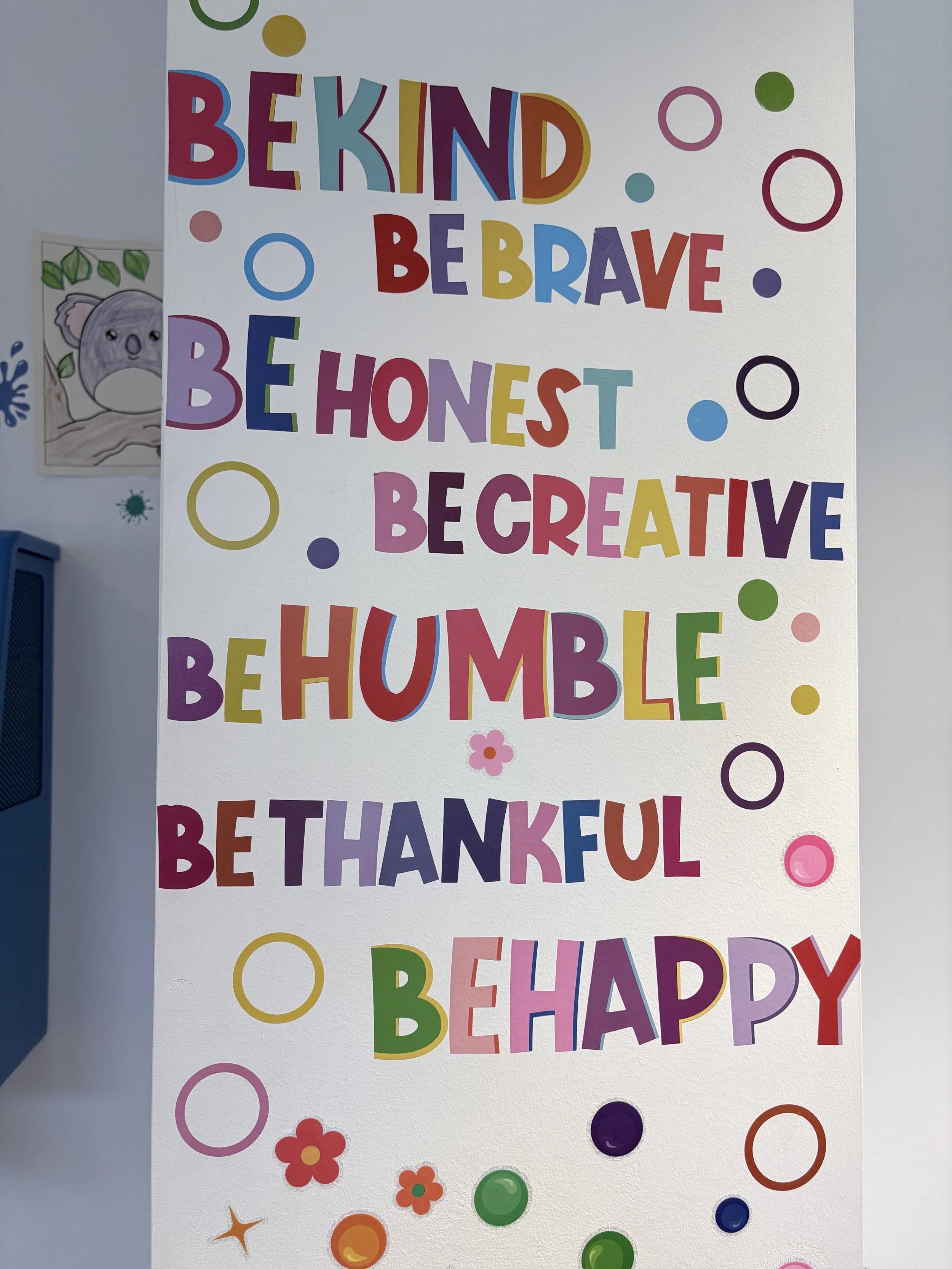 Colorful bulletin board with a message in multicolored letter cutouts reading, 'BE KIND, BE BRAVE, BE HONEST, BE CREATIVE, BE HUMBLE, BE HAPPY,' decorated with various circles, flowers, and stars.