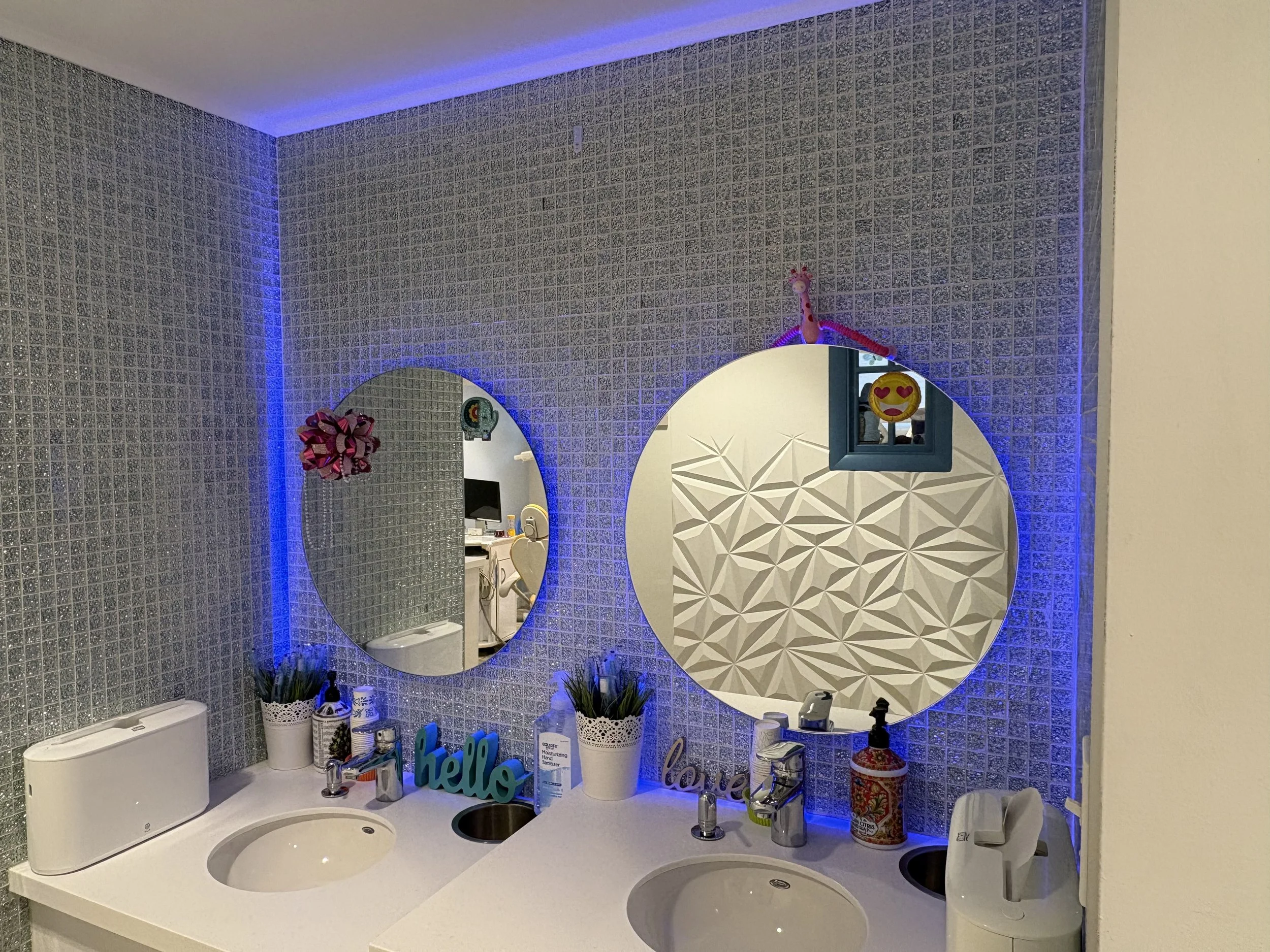 Bathroom vanity with two sinks, mirrors, and bathroom decor including a pink ribbon bow, emoji plush, and 'hello' word sign, illuminated by blue LED lighting.