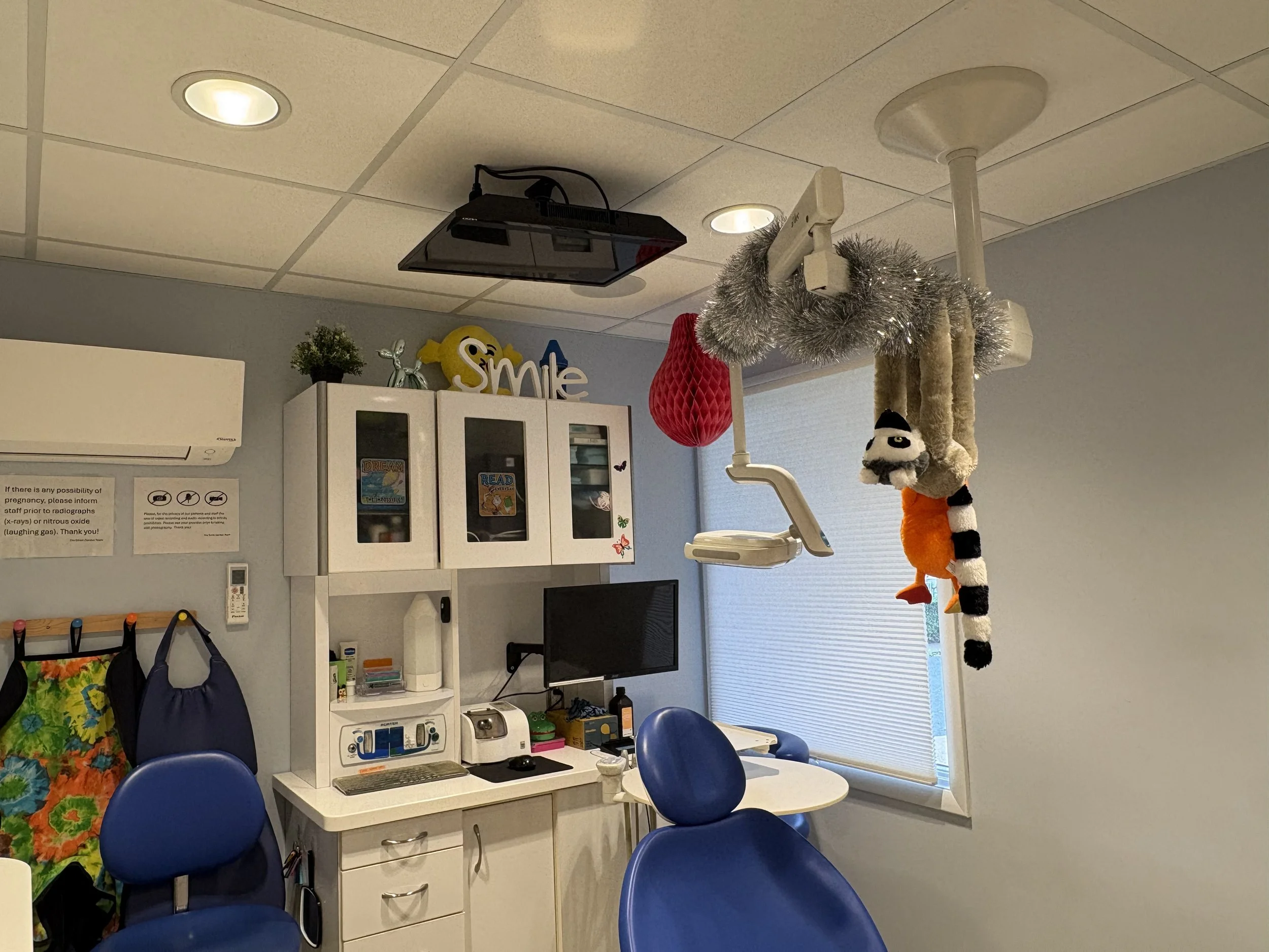 Hospital room with ceiling lights, medical equipment, a monitor, a white cabinet with books and decor, empty blue chairs, and hanging plush toys, including a panda and a clownfish.