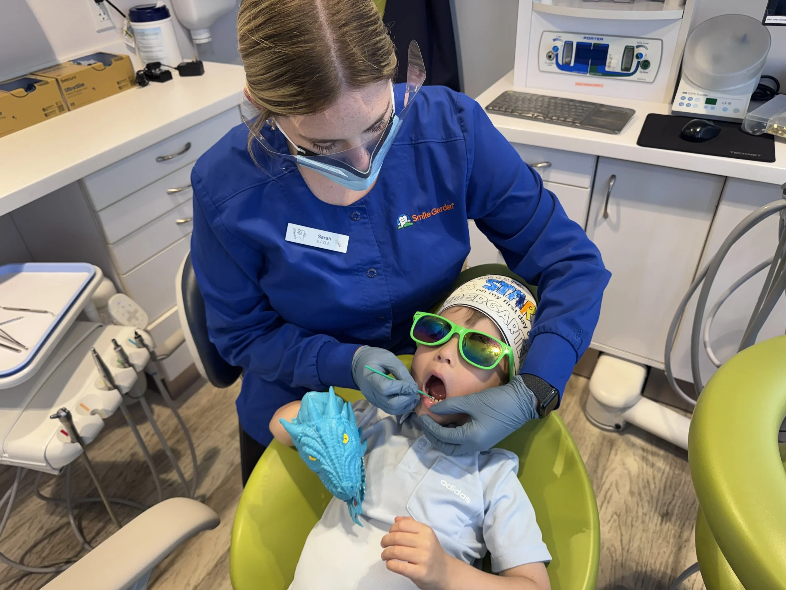 A child lying in a dental chair with sunglasses and a headband, receiving dental care from a dentist wearing protective glasses, a face mask, and gloves in a dental office.