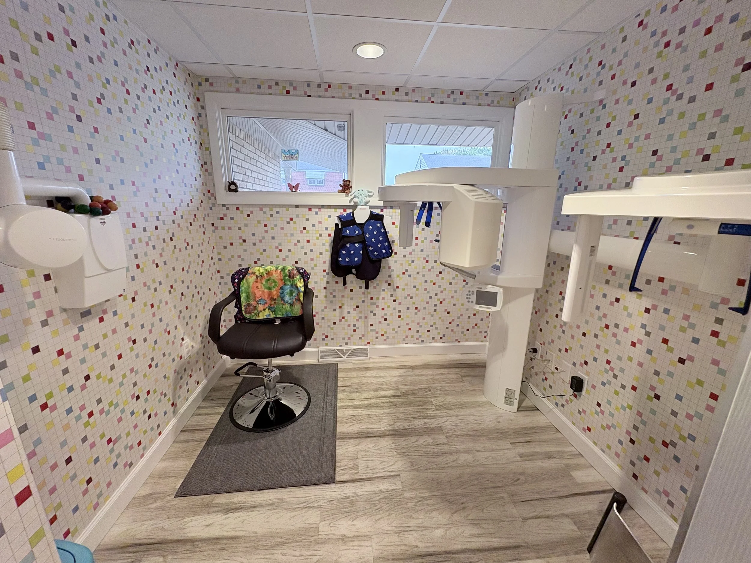 A small medical or dental examination room with colorful mosaic tile walls, a window with a view of the outside, a black rolling chair with a floral cushion, a white wall-mounted hand sanitizer dispenser with colorful candies on top, a white dental or medical equipment stand, a small gray rug, a gray flooring, and a shelving unit or cabinet on the right side.