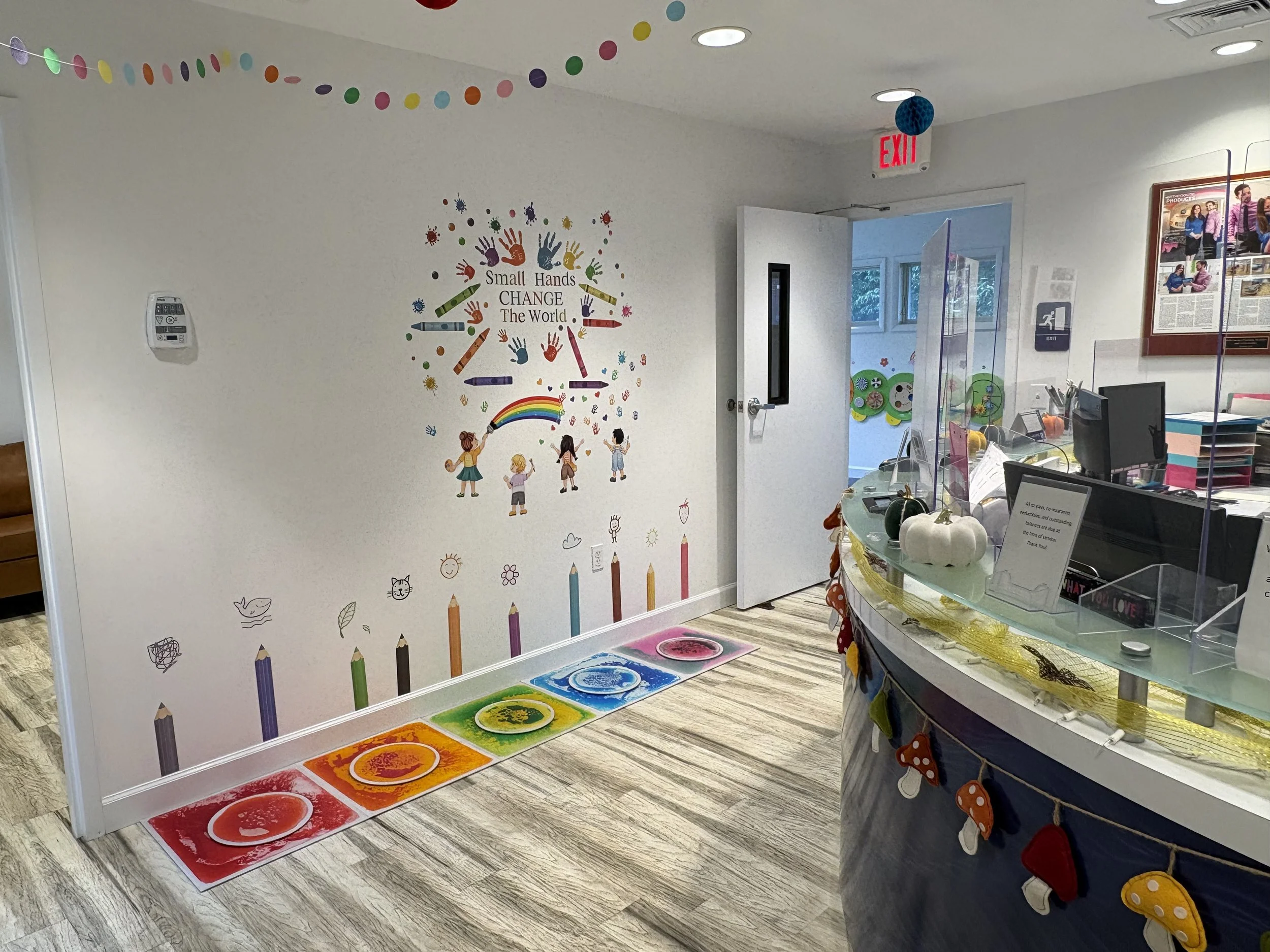 Colorful children's artwork and decorations inside a community center or classroom. A wall features a rainbow, children holding a rainbow hose, and colorful handprints with the message 'Small Hands Change The World.' The floor has a rainbow-colored m