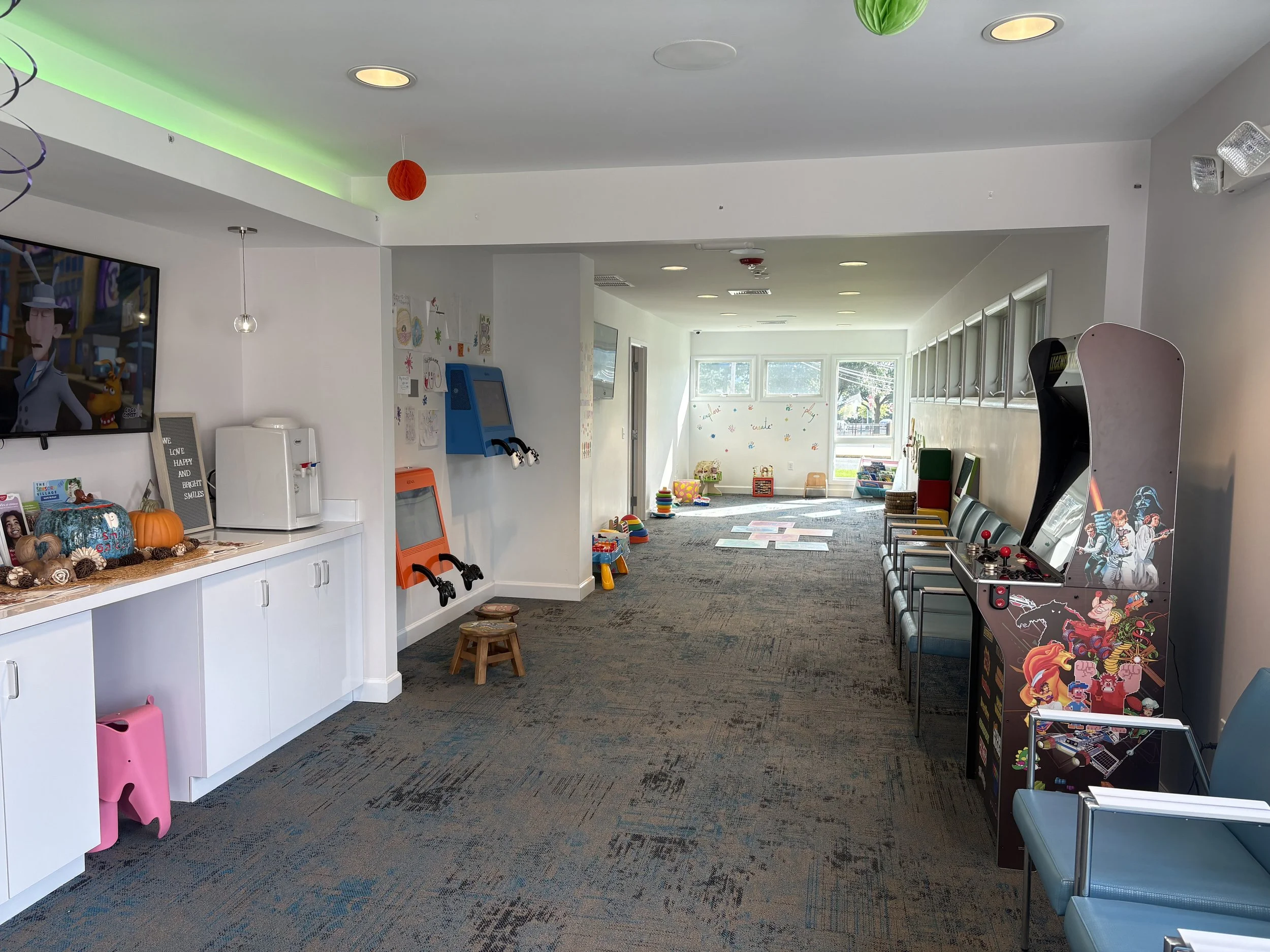 Interior of a children's play or activity area, decorated with Halloween pumpkins, gourds, and autumn-themed decor on a white counter. There are colorful toys, a small orange and gray art easel, and arcade-style gaming machines along the walls. The room is bright with natural light from windows at the far end, and children's artwork displayed on the wall. The ceiling has sports balls hanging as decoration.
