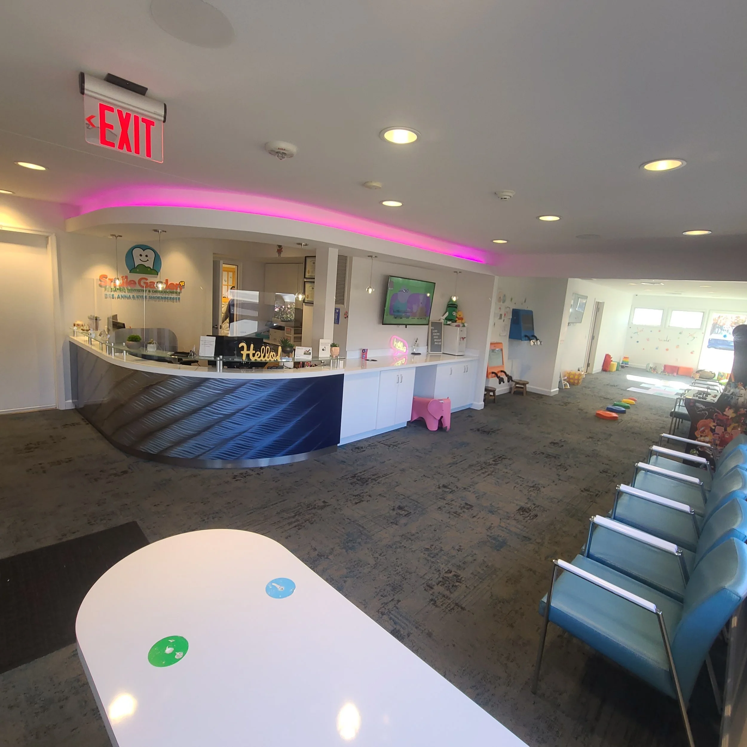 Reception area of a dental office with a white counter, signage, and colorful decor, including a cartoon tooth logo on the wall, a television screen, and children's toys on the floor.