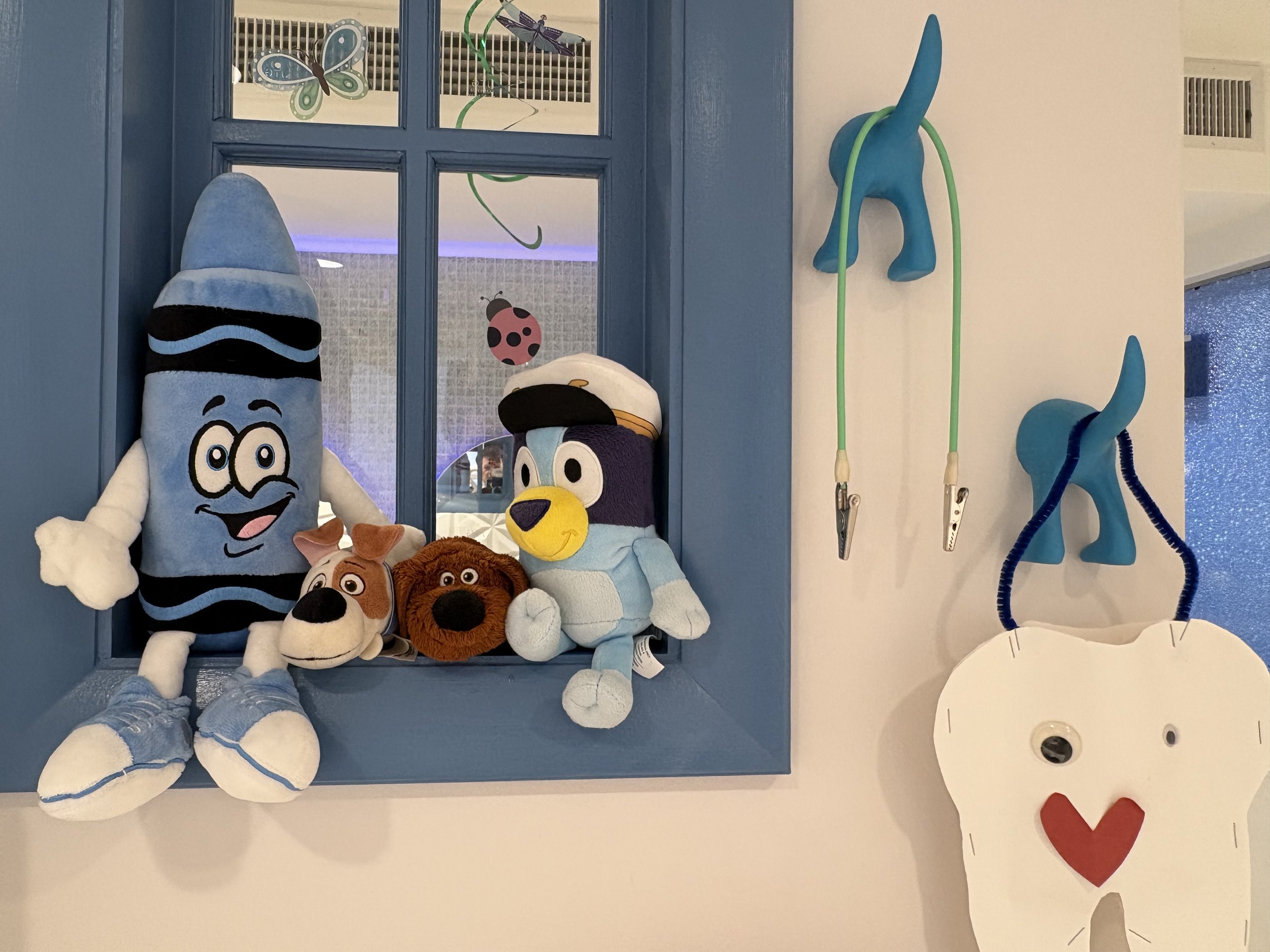Various plush toys including a blue sock monkey, a dog, a penguin, and a bear sitting on a blue windowsill. Behind the toys, a window with decorative butterfly and dragonfly stickers is visible. To the right, there are blue hooks shaped like animals 