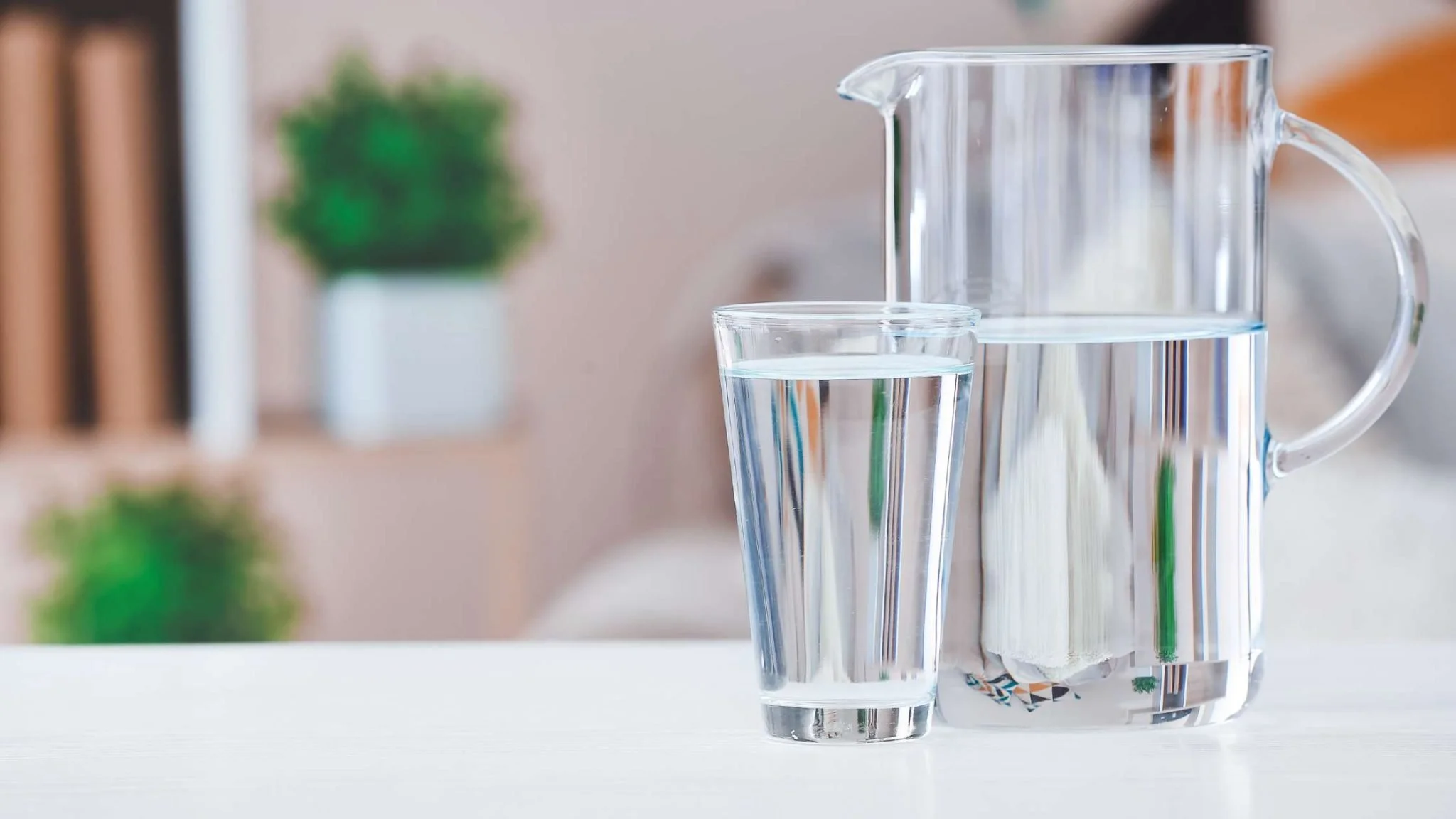 Clear glass pitcher and two glasses on a white surface, with a blurred background including a green plant.