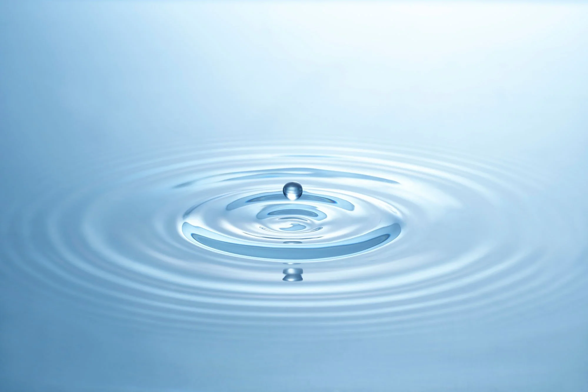 A close-up of a single water droplet falling into a calm water surface, creating ripples.