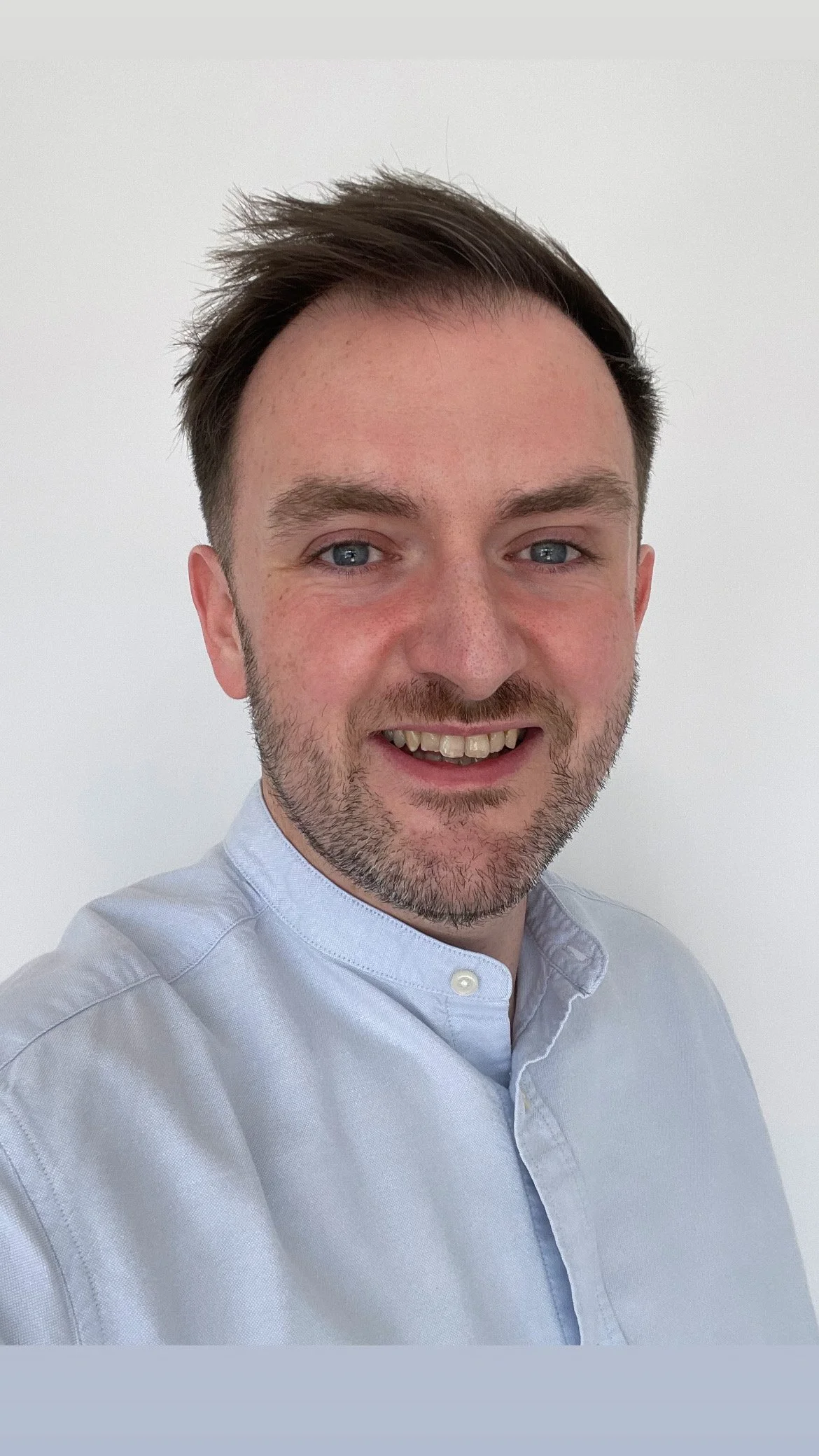 A smiling man with short brown hair and blue eyes, wearing a light blue button-up shirt, standing against a plain white background.