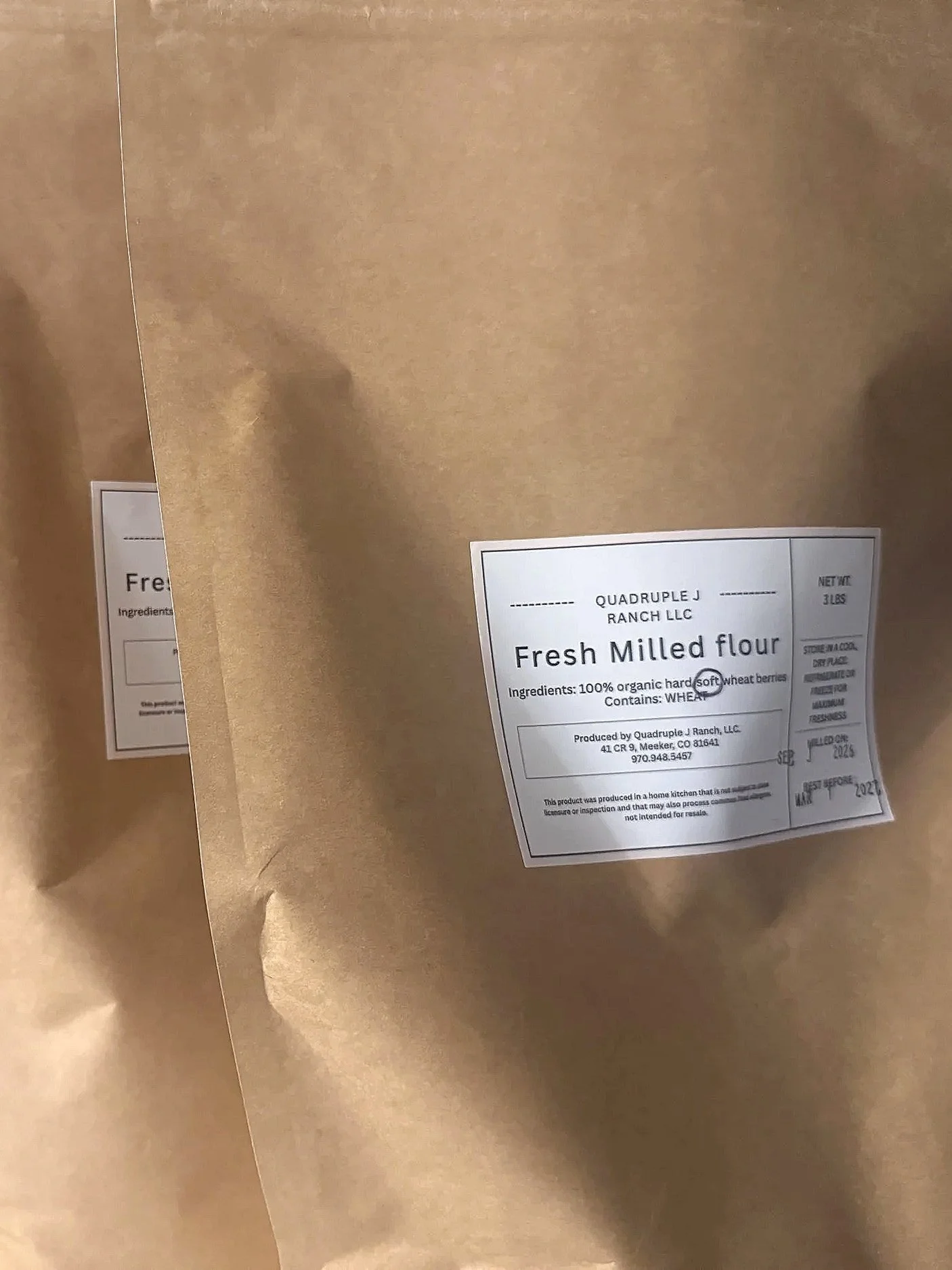 Brown paper bag of fresh milled flour with a label indicating it is from Quadruple J Ranch LLC, containing 100% organic hard soft wheat berries, produced in Mesker, Colorado.