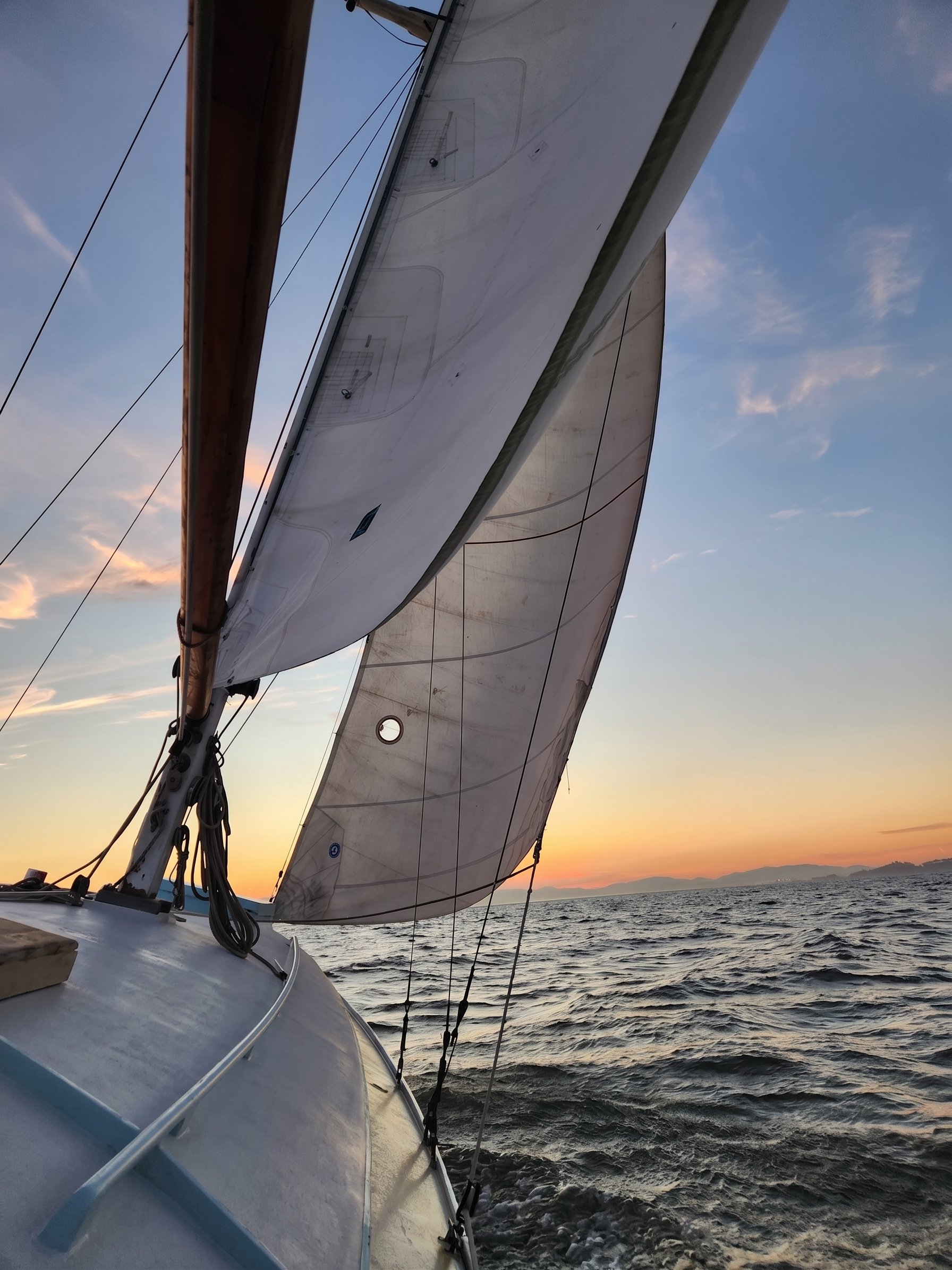 View from a sailboat at sunset, showing sails and part of the boat, with water and distant mountains in the background.