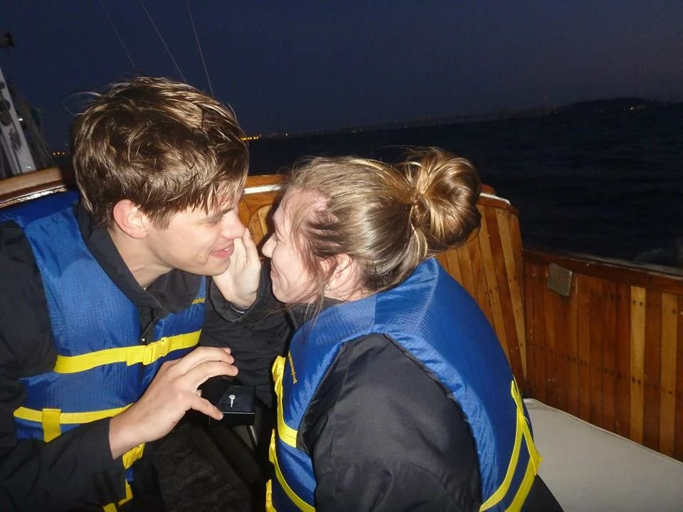 A young man and woman with life jackets on are close, leaning in for a kiss or whispering to each other on a boat at night.