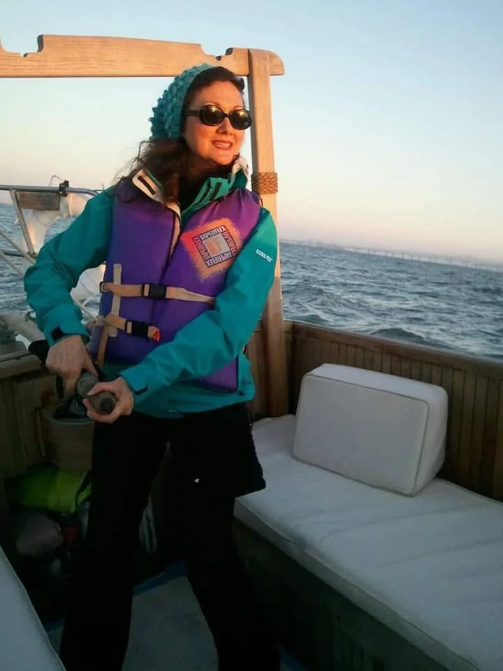 Woman wearing sunglasses, a blue knit hat, a colorful life jacket, and a teal jacket standing on a boat, holding a steering wheel with the ocean visible in the background during sunset.
