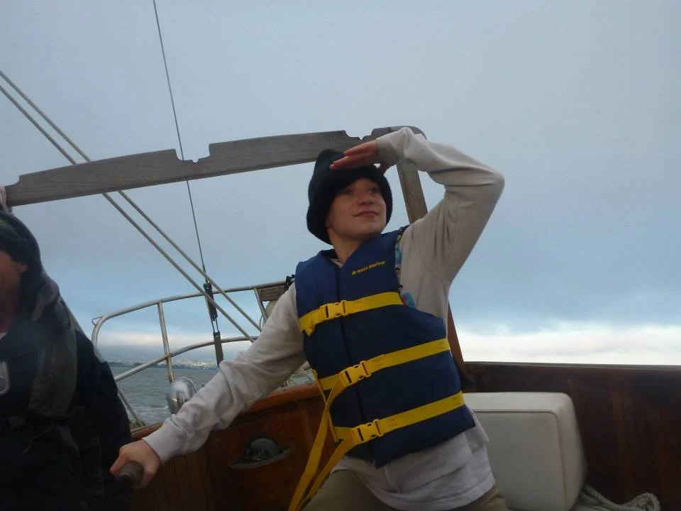 Person on a boat, wearing a life vest and a black beanie, holding onto the boat's railing and shading eyes while looking into the distance, with stormy skies overhead.