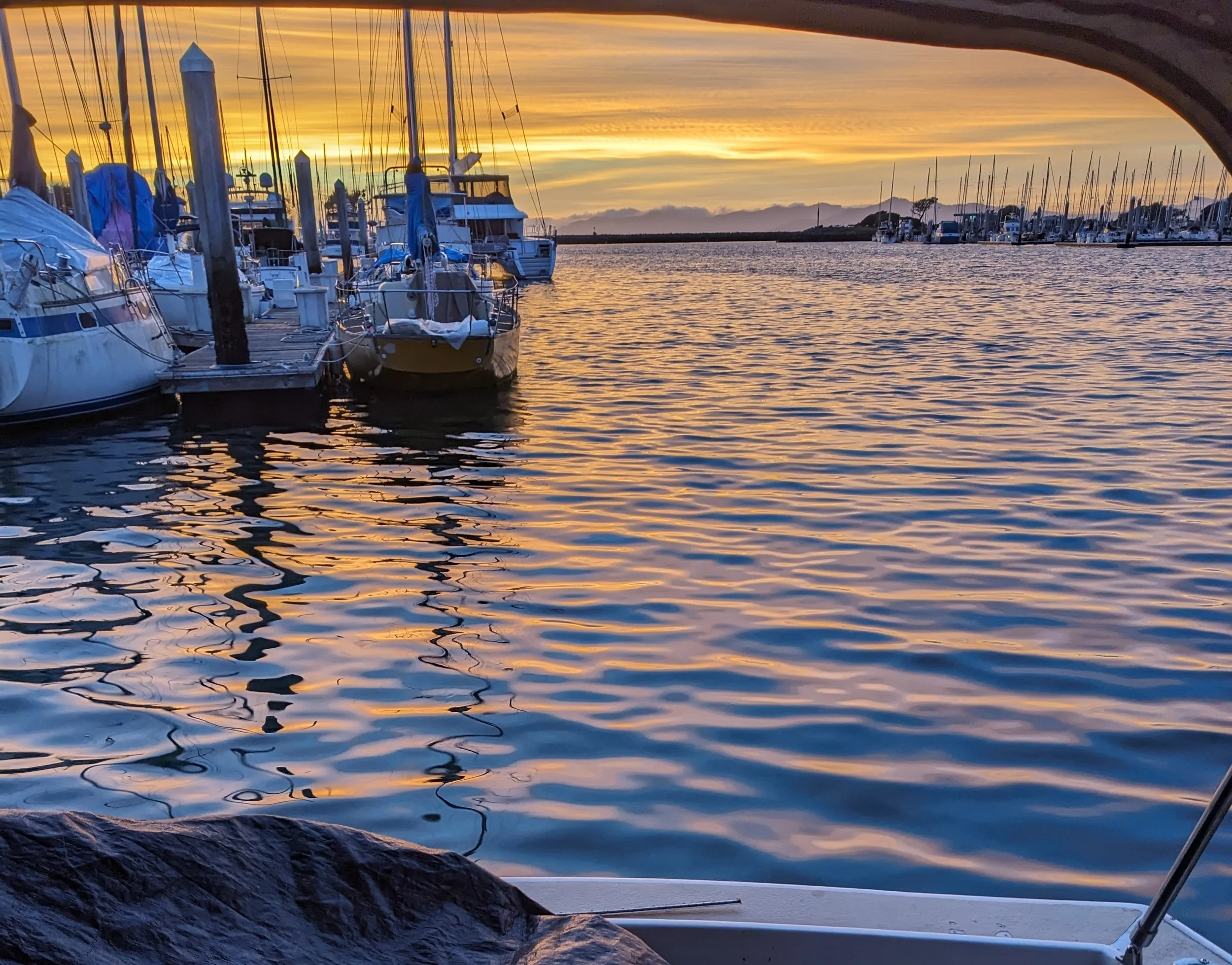 Sunset over a marina with boats docked, reflecting vibrant orange and yellow hues on the water, framed by a boat's bow and railing in the foreground.