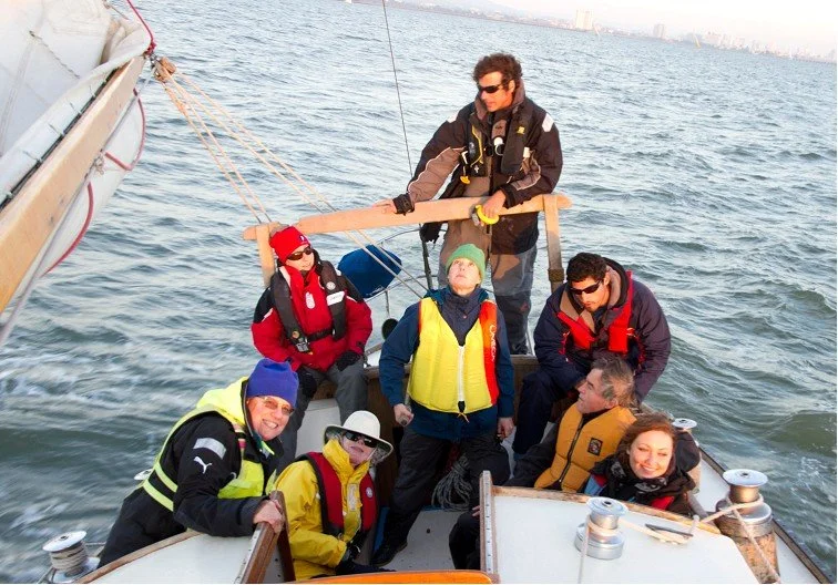 Group of seven people on a boat, wearing life jackets and outdoor gear, some with sunglasses and hats, enjoying a day on the water.