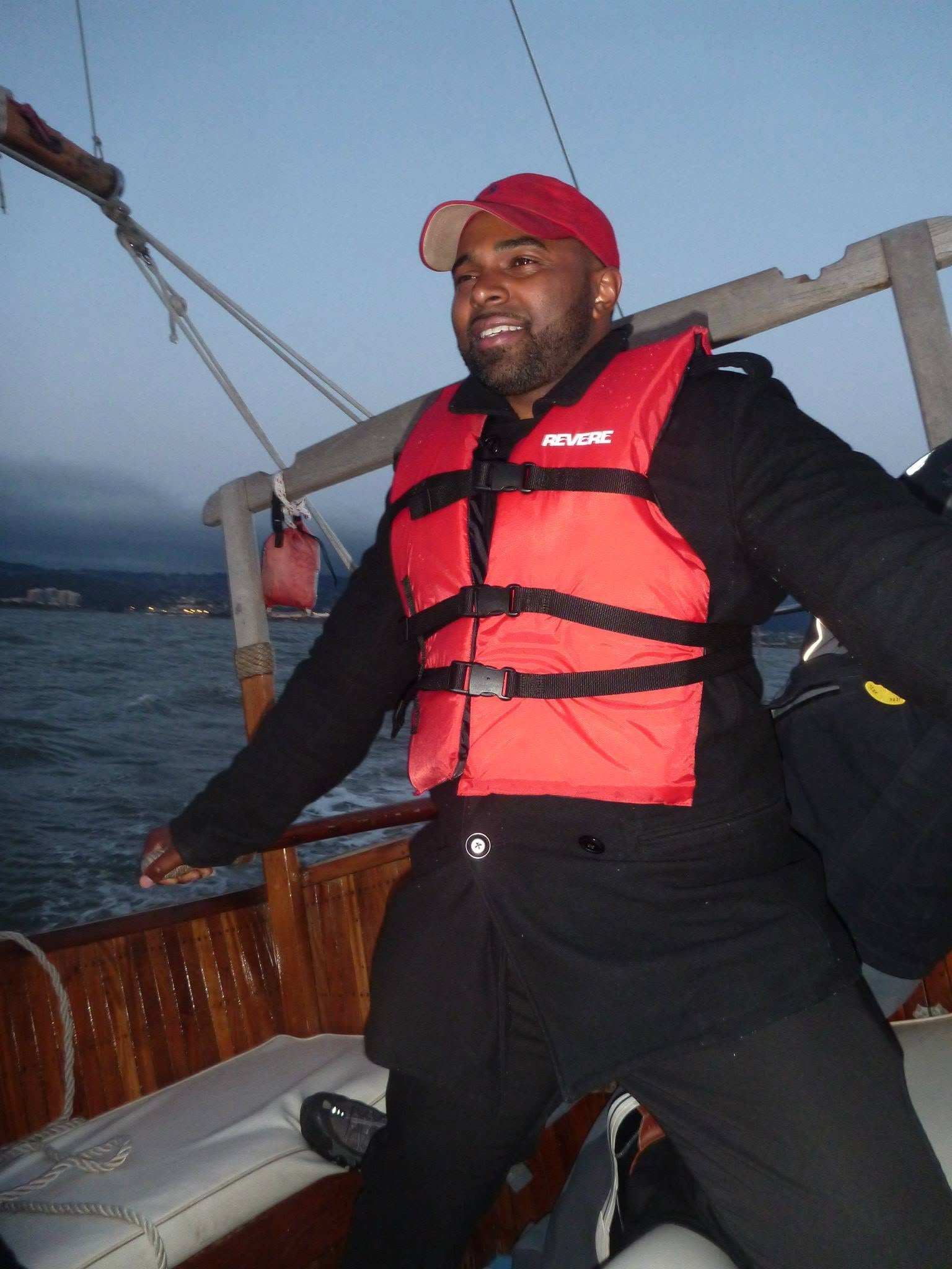A man in a red hat and life jacket on a boat, with water and a distant shoreline in the background.