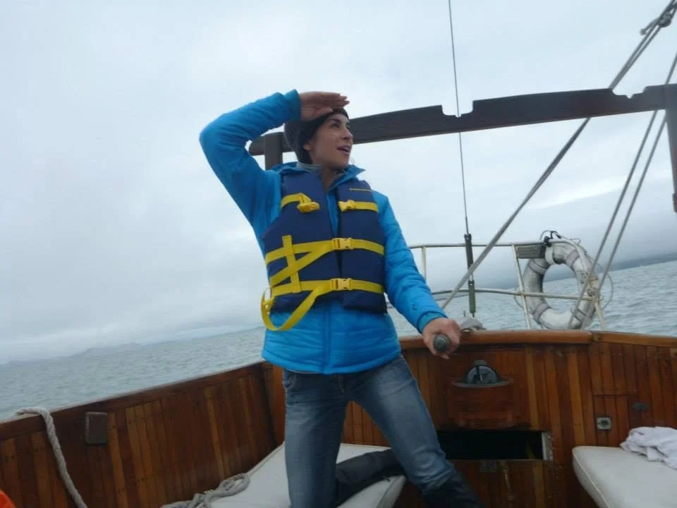Woman wearing a blue jacket and life vest standing on a boat, looking into the distance with her hand on her forehead.