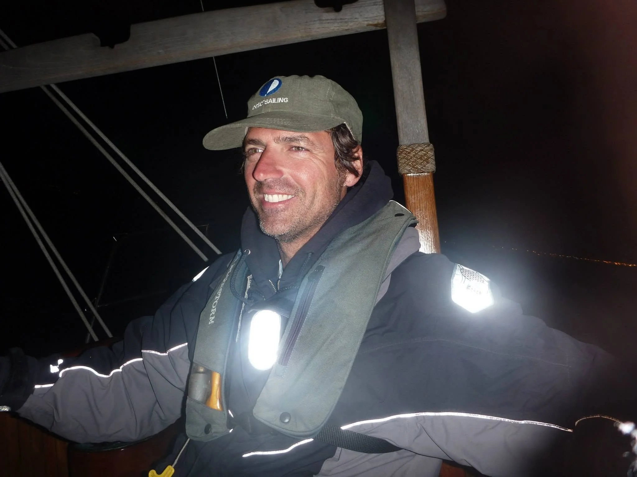 Captain Dan wearing a cap with 'Pacific Sailing' written on it, smiling at night with a dark sky in the background, leaning on a wooden railing aboard a sailboat. Reflective safety stripes glow from the camera flash.