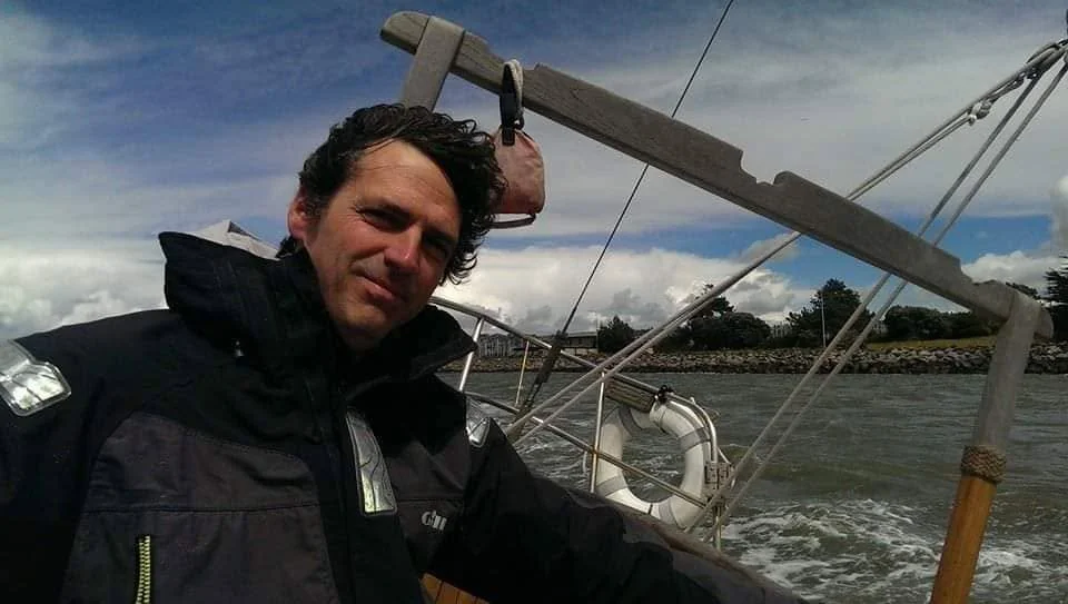 A man on a sailboat holding the tiller, with a cloudy sky and shoreline in the background.