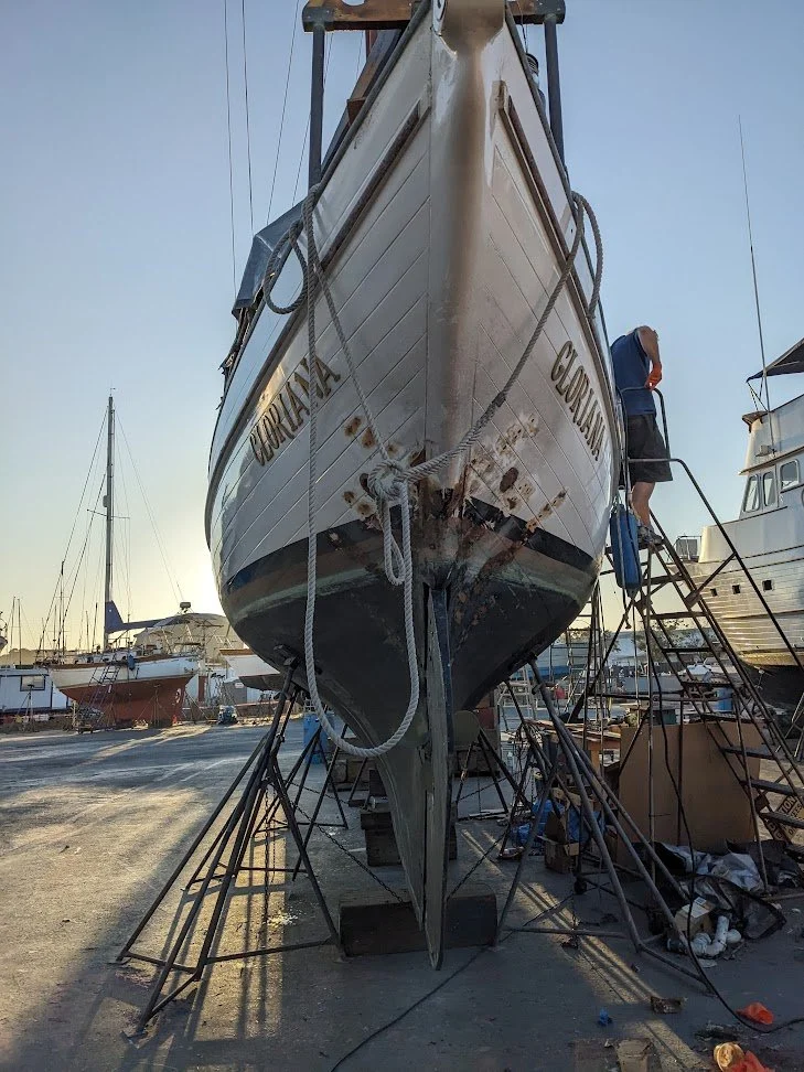 A boat named 'GLORIANA' is on dry dock, with a person working on it. The boat is propped up on stands in a boatyard with other boats visible in the background.
