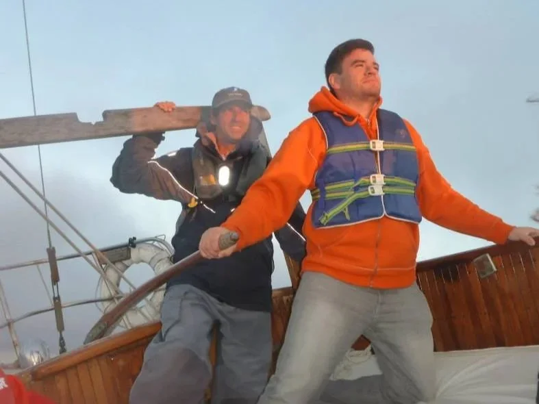 Two men stand on a boat. Captain Dan stands behind a client, who has taken the tiller. Both men are smiling and looking forward towards the horizon.