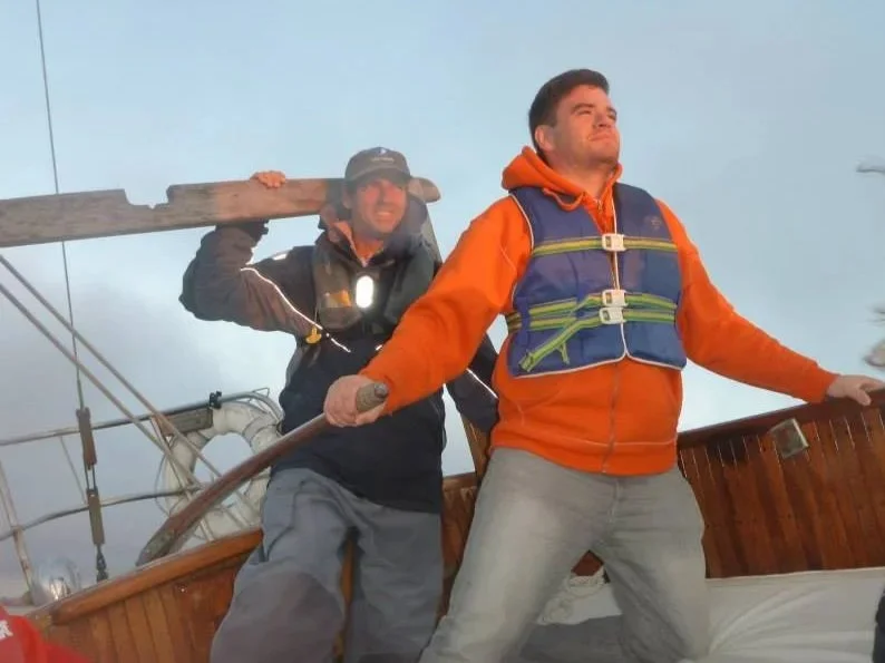 Two men on a boat, one holding a wooden plank, the other gripping a rope, with cloudy sky in the background.