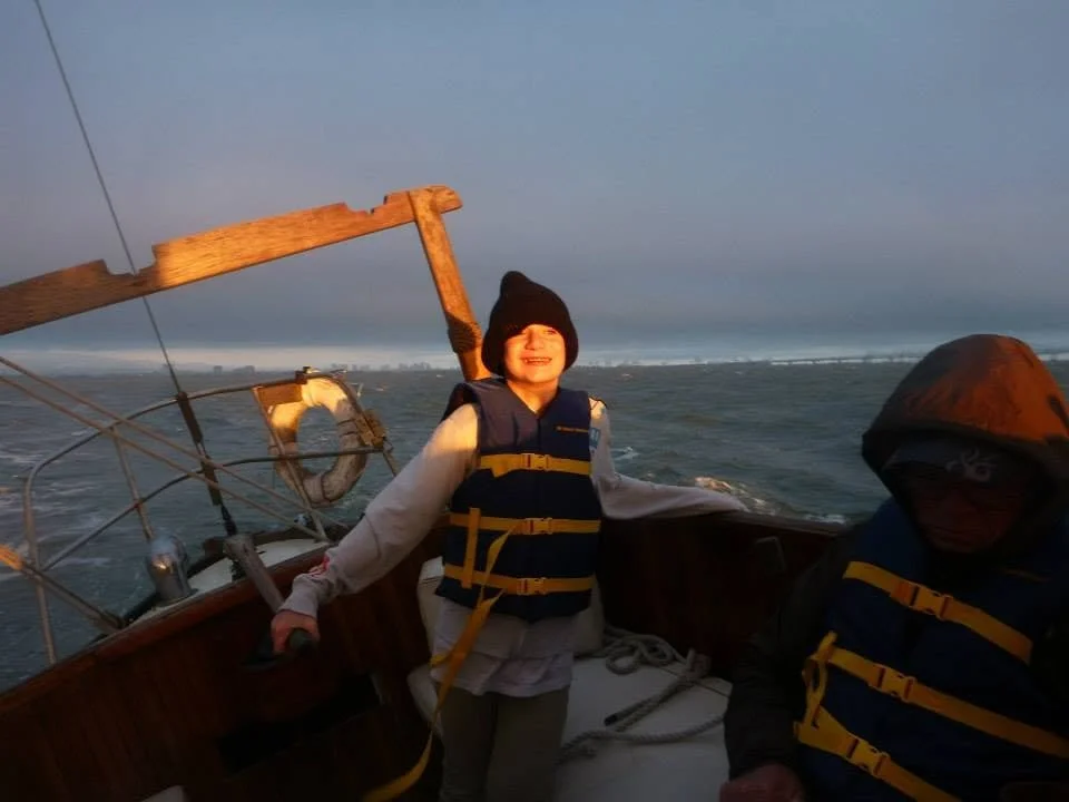 A smiling child and an adult on a boat, wearing life jackets, with large water waves and a cloudy sky in the background.
