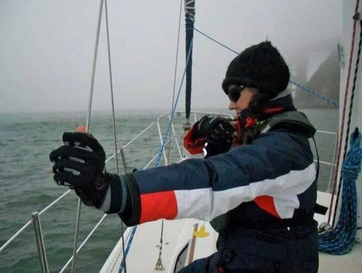 A person dressed in a black beanie, sunglasses, and a navy, white, and red jacket, took a selfie on a sailboat during foggy weather.