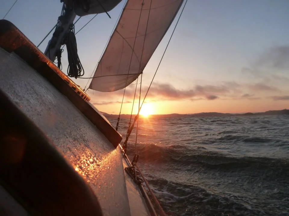 View from a sailboat during sunset, showing the boat's hull, sail, and the water with waves.