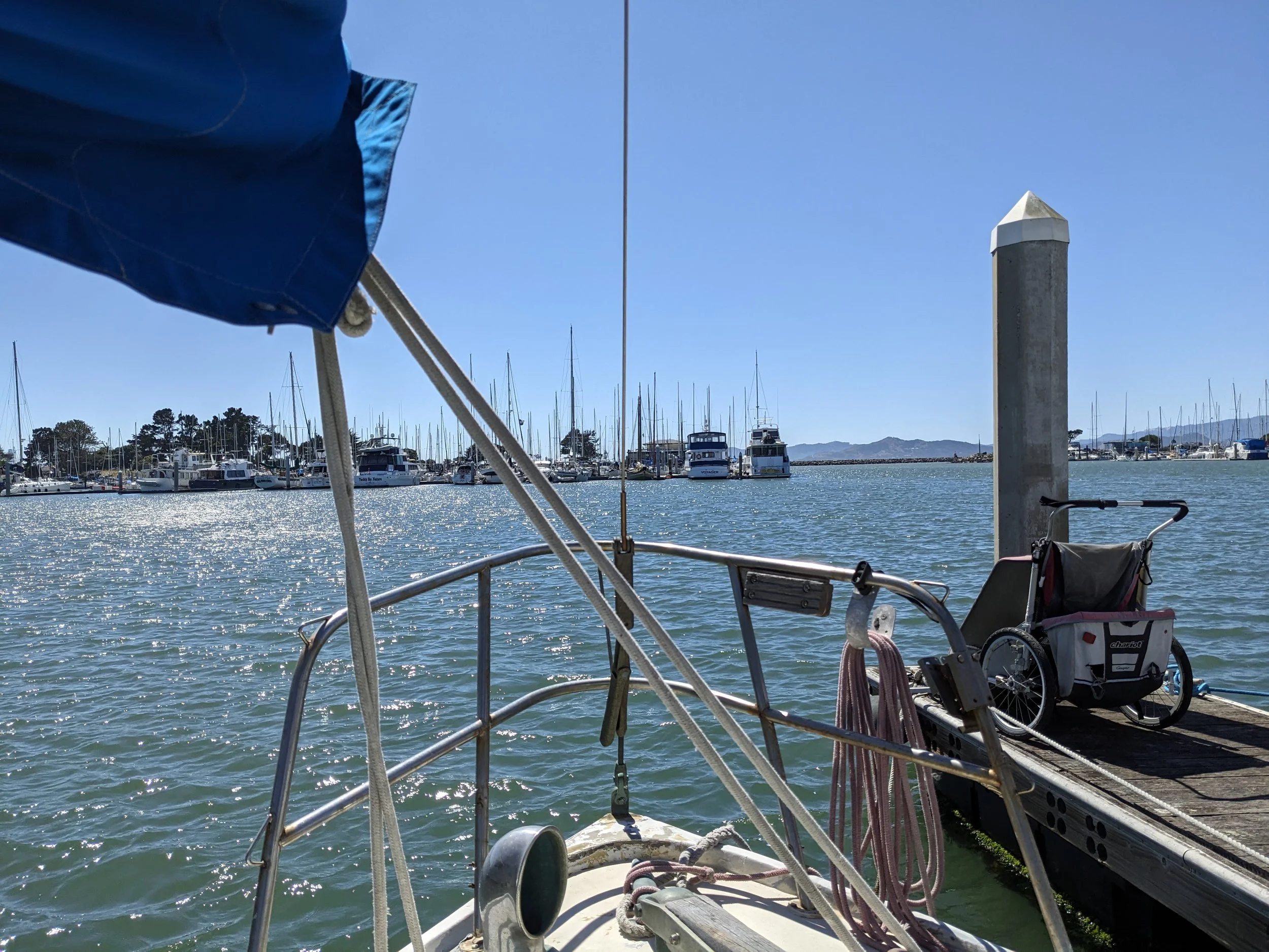 View from a boat dock showing a marina with numerous sailboats and yachts anchored in the water, a large white post, and mountains in the distance under a clear blue sky.
