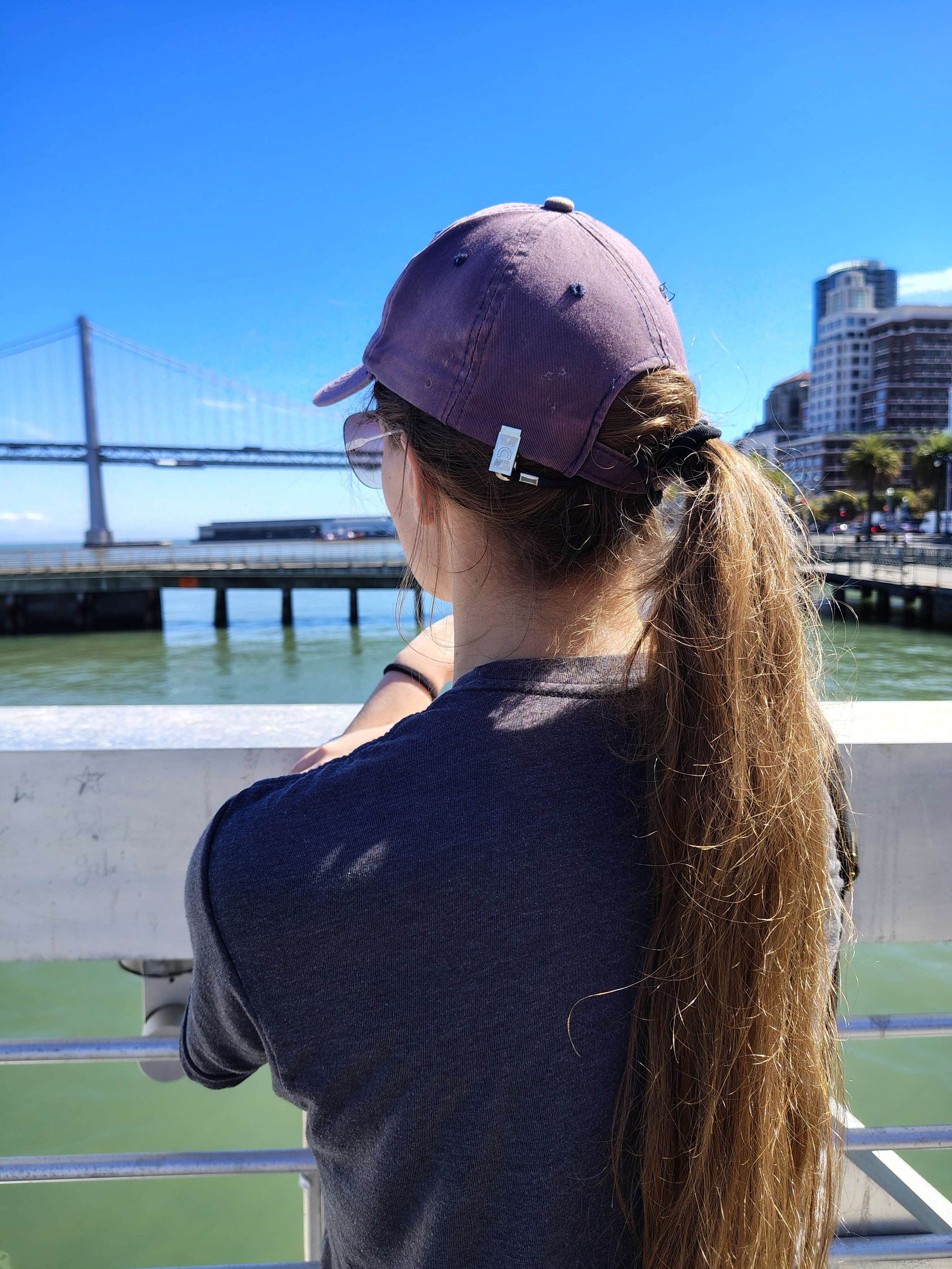 A woman with long hair wearing a purple baseball cap and sunglasses, standing on a boat or ferry, looking at a bridge over water with city buildings in the background on a clear, sunny day.