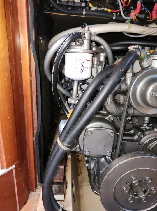 Close-up of an engine compartment showing various hoses, cylinders, and mechanical components, including a white oil filter with handwritten notes.