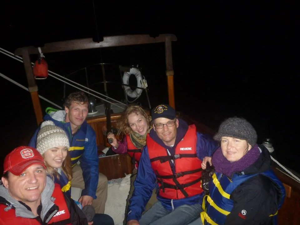 Group of six people on a boat at night, smiling and dressed for cold weather with life jackets and hats.