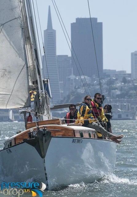 Sailing boat with four people on water, San Francisco skyline in background.