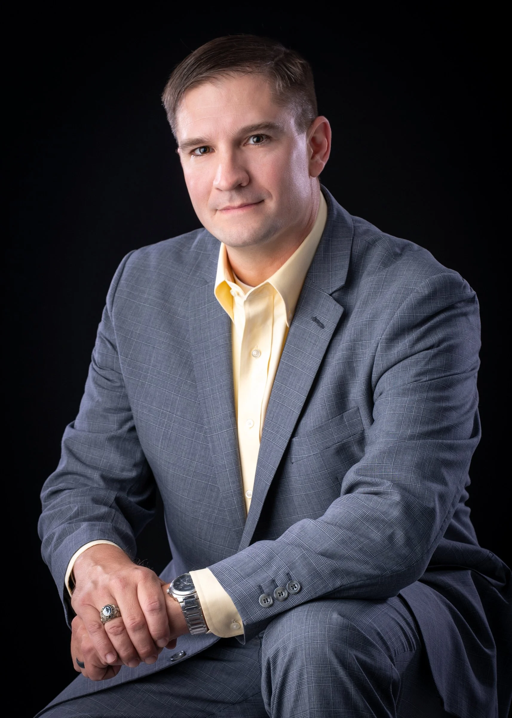 A young man in a gray plaid suit with a yellow shirt, sitting against a black background, looking at the camera, with his hands clasped and wearing a watch and rings.