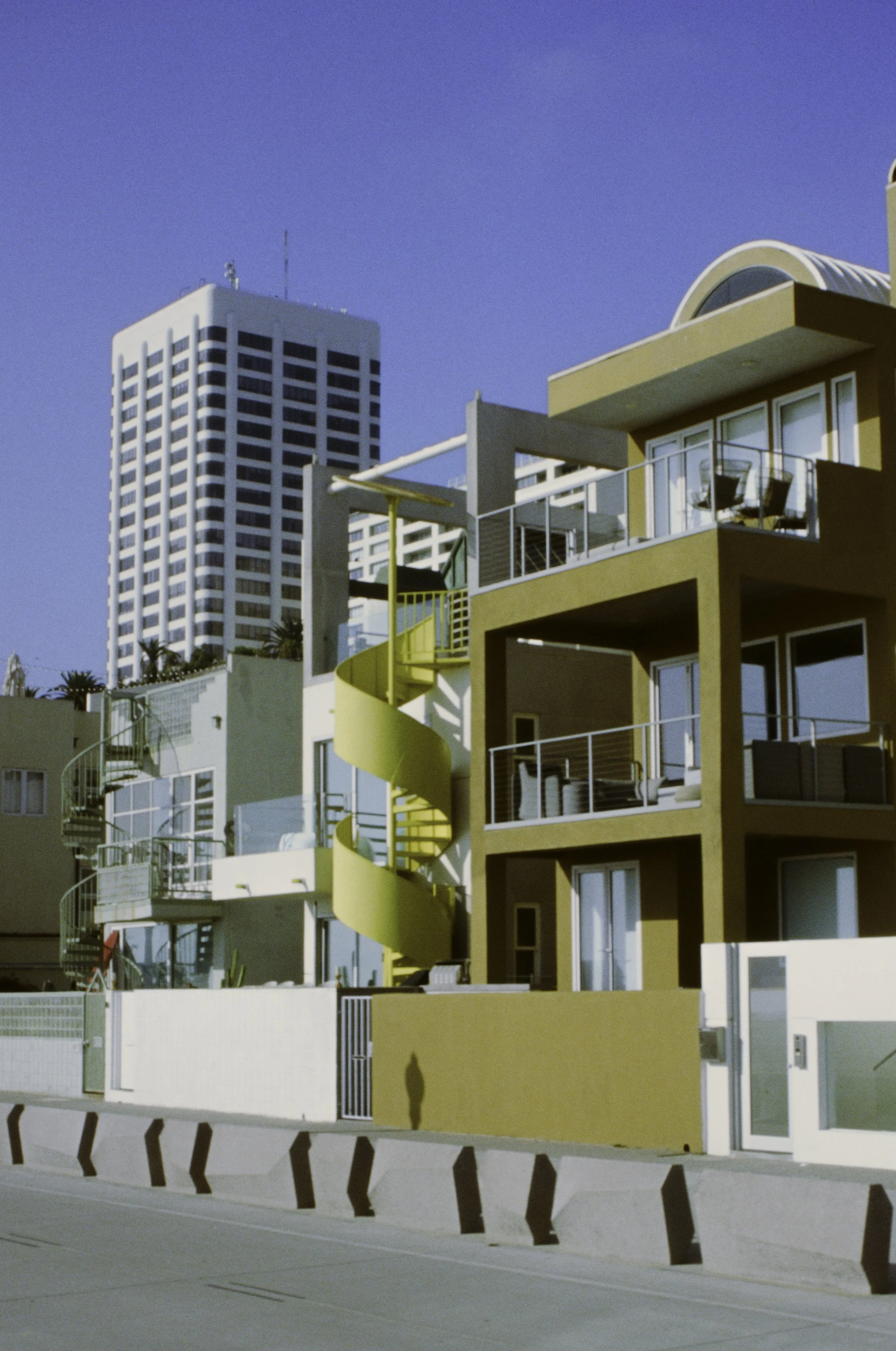 Colorful modern apartment building with yellow spiral staircase on balcony, set against a clear blue sky, with a tall white office building in the background.
