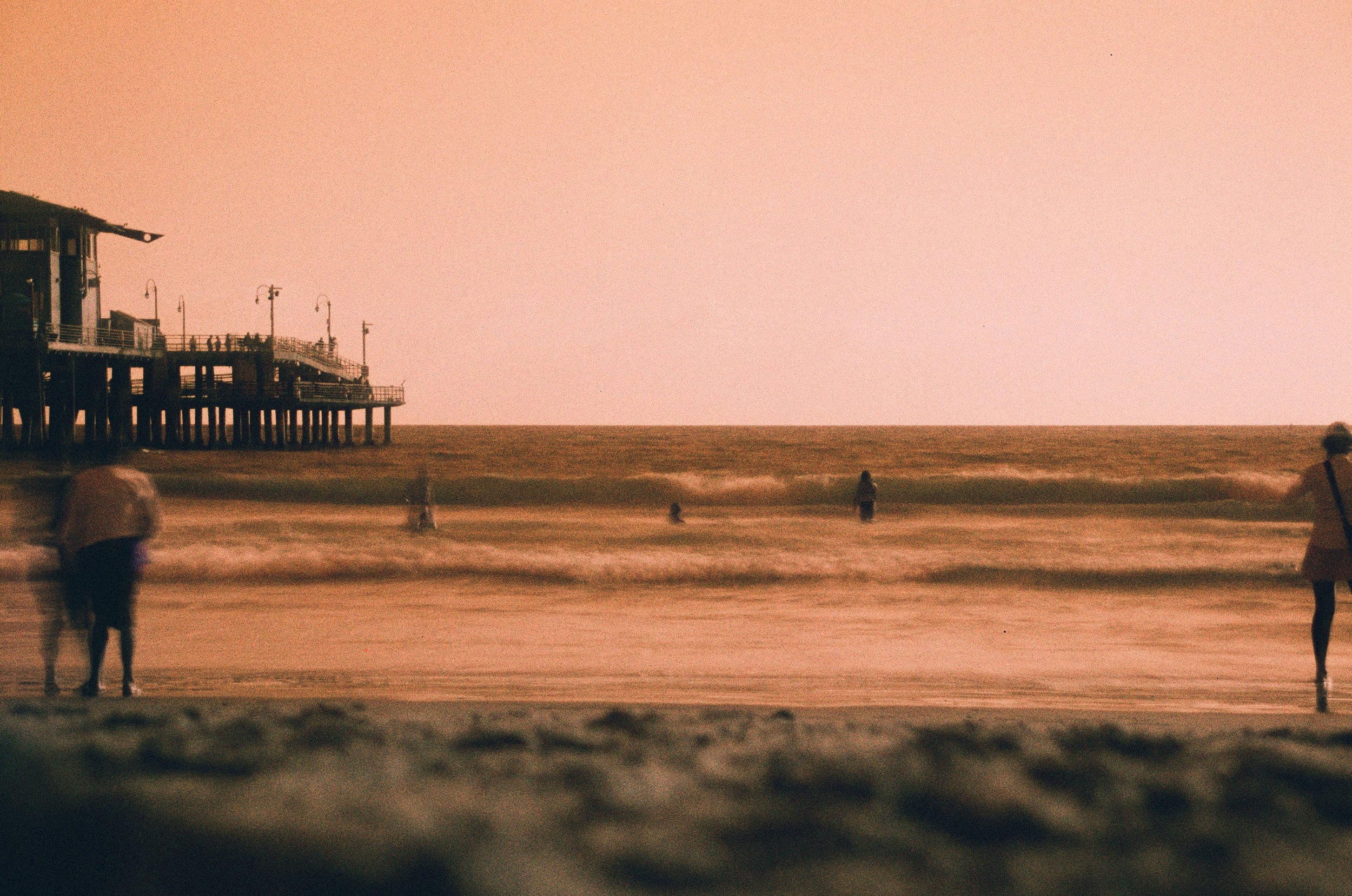 Beach scene at sunset with a pier on the left, people walking along the shore, and a pinkish sky.