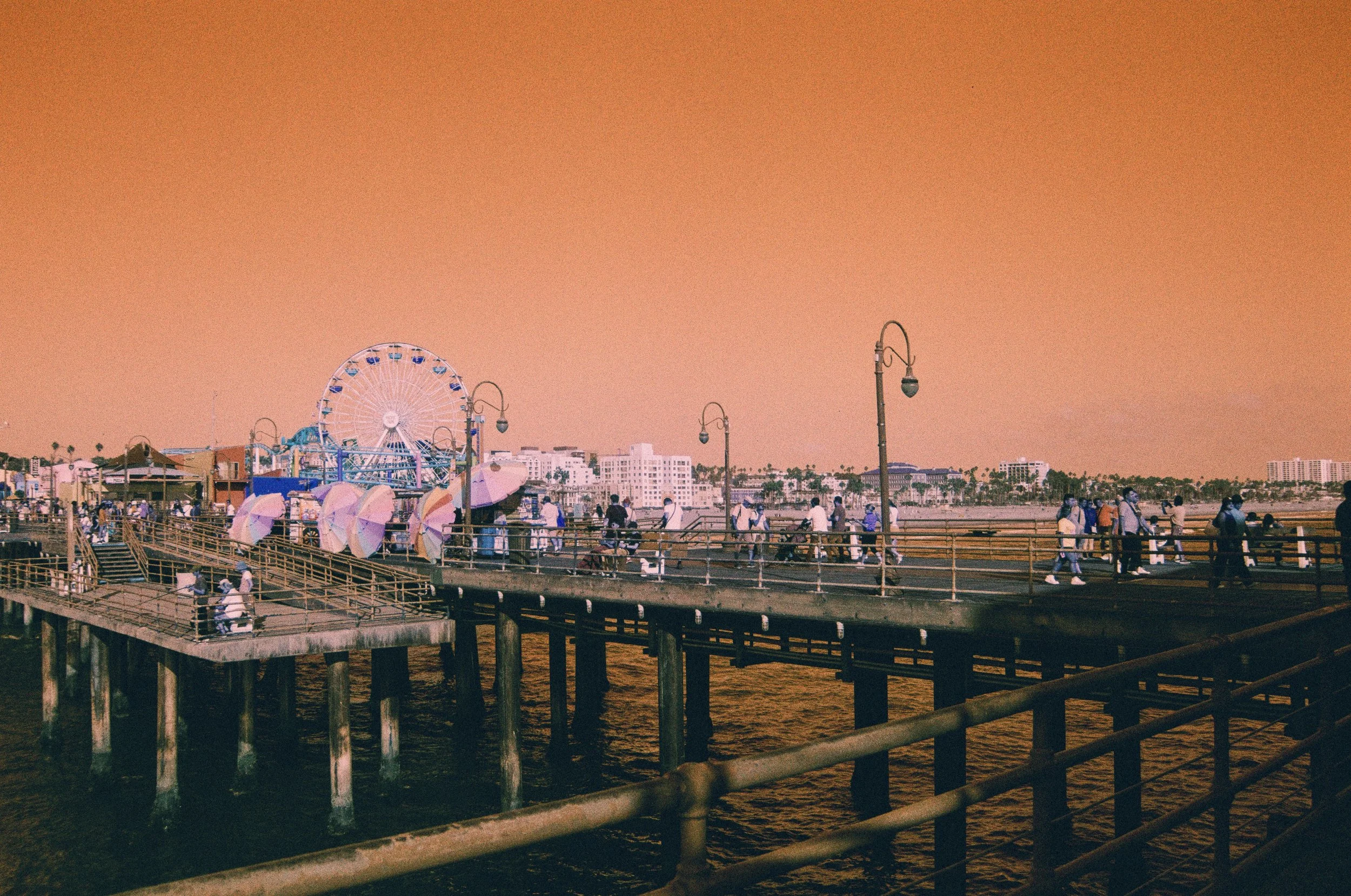 People walking and biking on a pier at sunset, with a Ferris wheel and city buildings in the background.