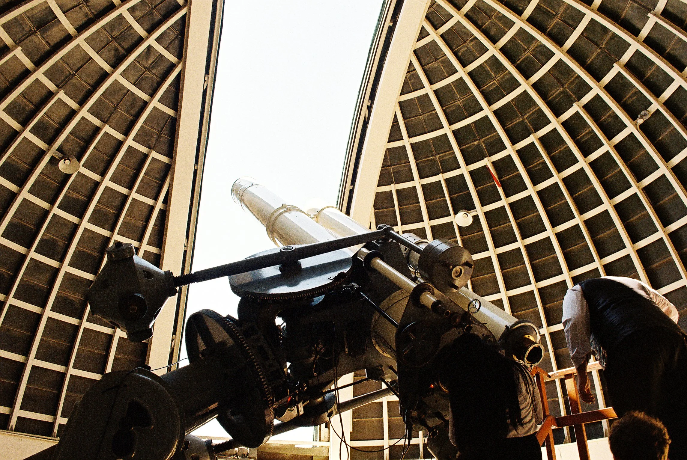 Looking up through the open roof of a building, a telescope is aimed upward, with a person adjusting the telescope in the foreground and another person nearby. The roof has a grid-like pattern, and the sky is visible above.