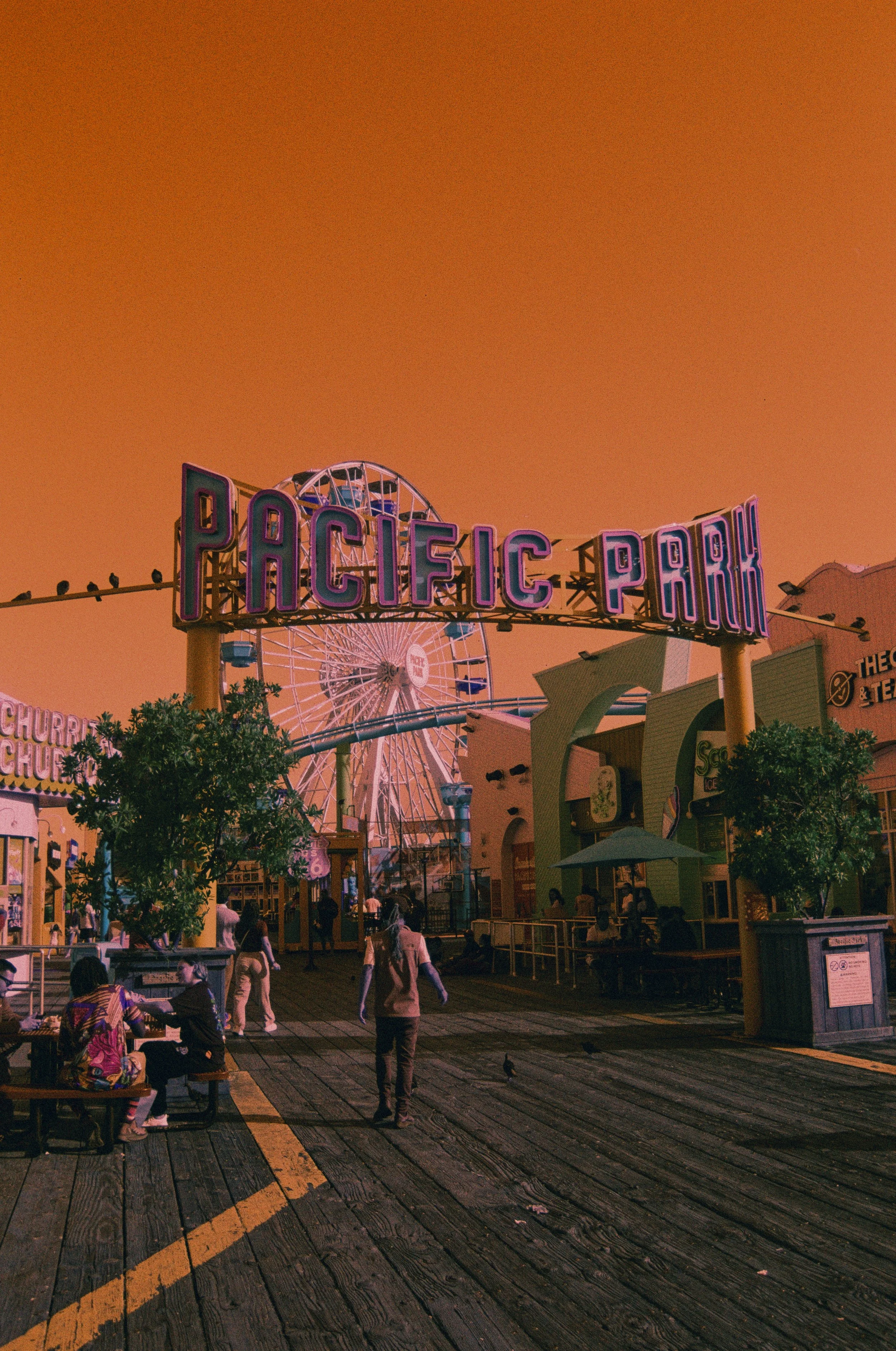 A lively amusement park scene at sunset with a large neon sign reading 'Pacific Park,' a Ferris wheel in the background, and people walking and sitting on benches in the foreground.