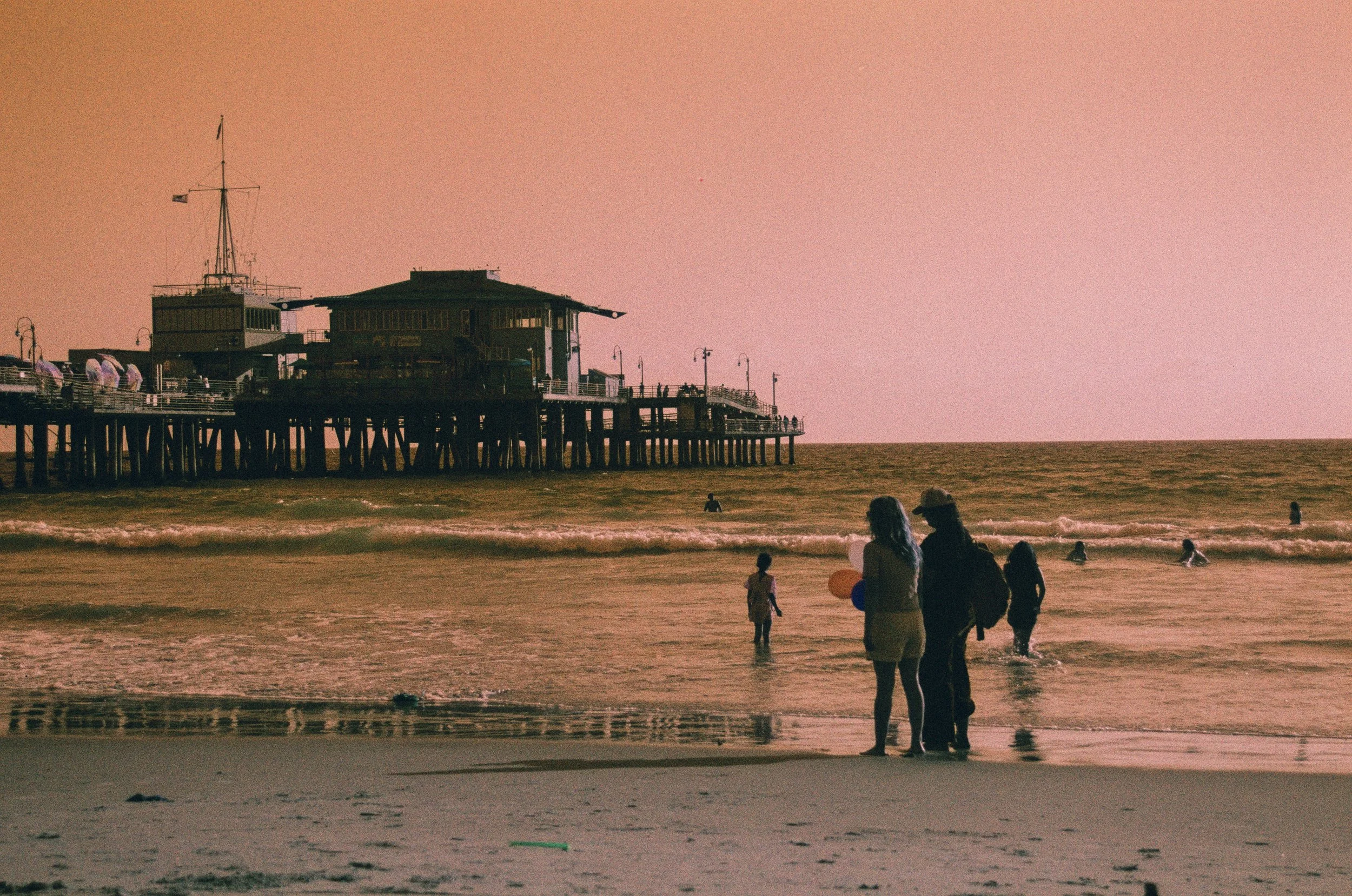 A pier extending into the ocean with a building on it, people on the beach, and some in the water during sunset or sunrise with a pink sky.
