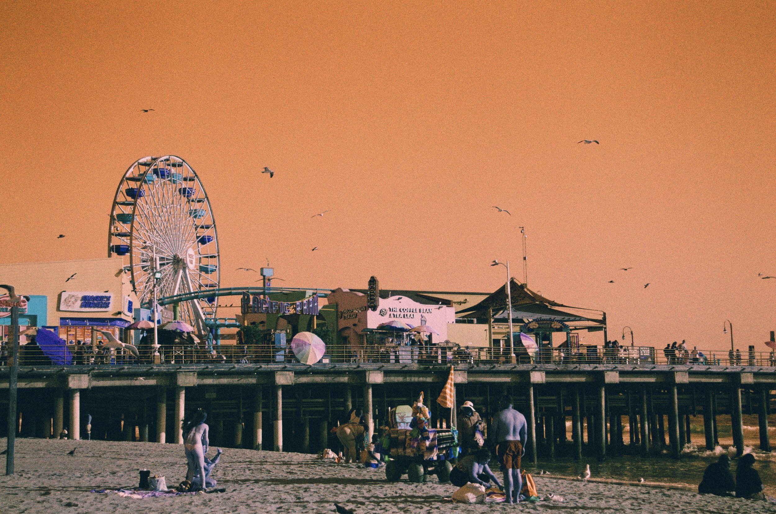 Beach scene at sunset with people walking and sitting on the sand, amusement park rides on a pier including a Ferris wheel, and seagulls flying in the sky.