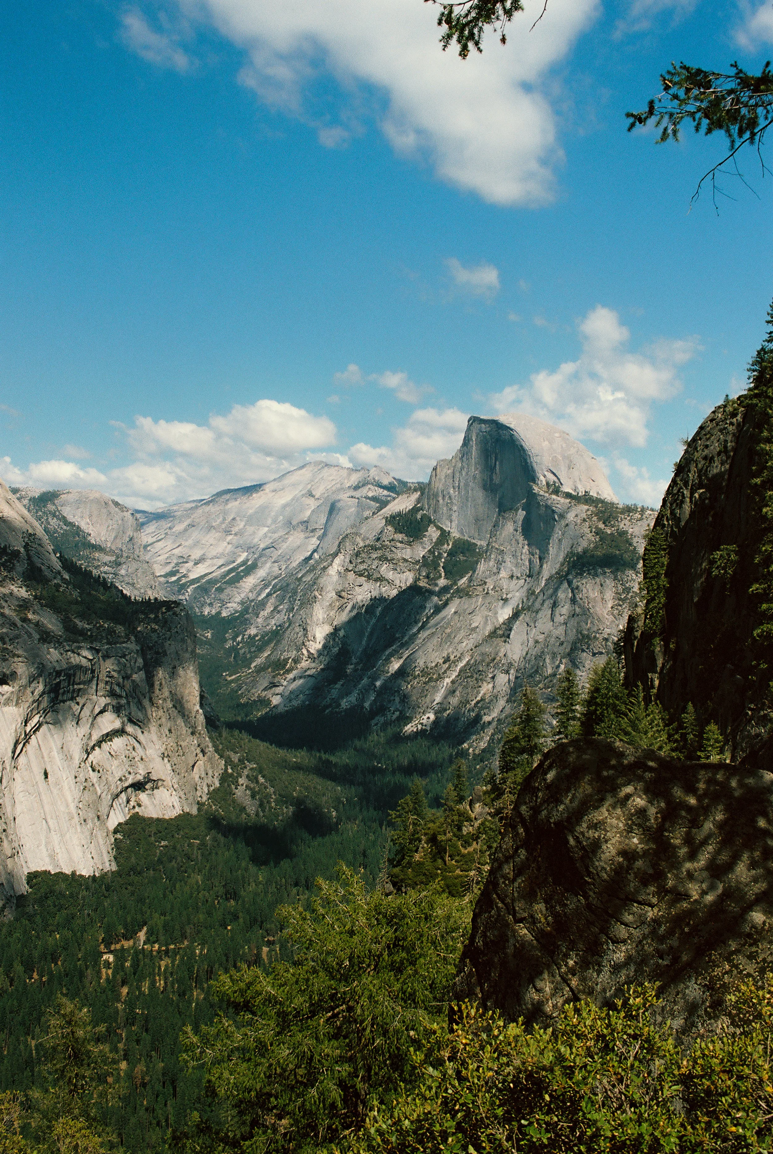A mountain landscape with large granite cliffs, green forested valley, and a partly cloudy sky.