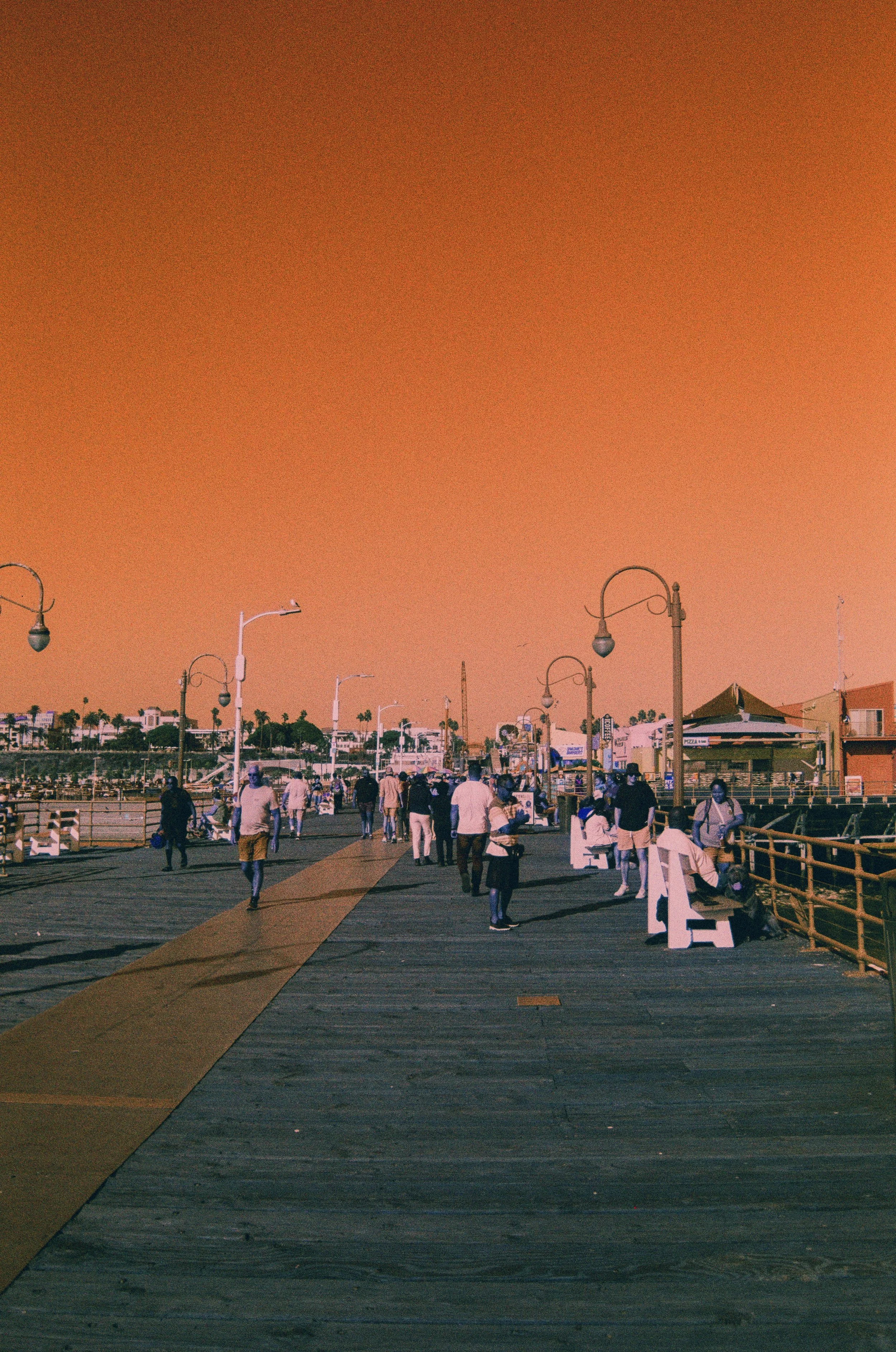A crowded wooden pier or boardwalk at sunset with people walking and socializing, under street lamps, leading towards buildings and boats in a harbor.